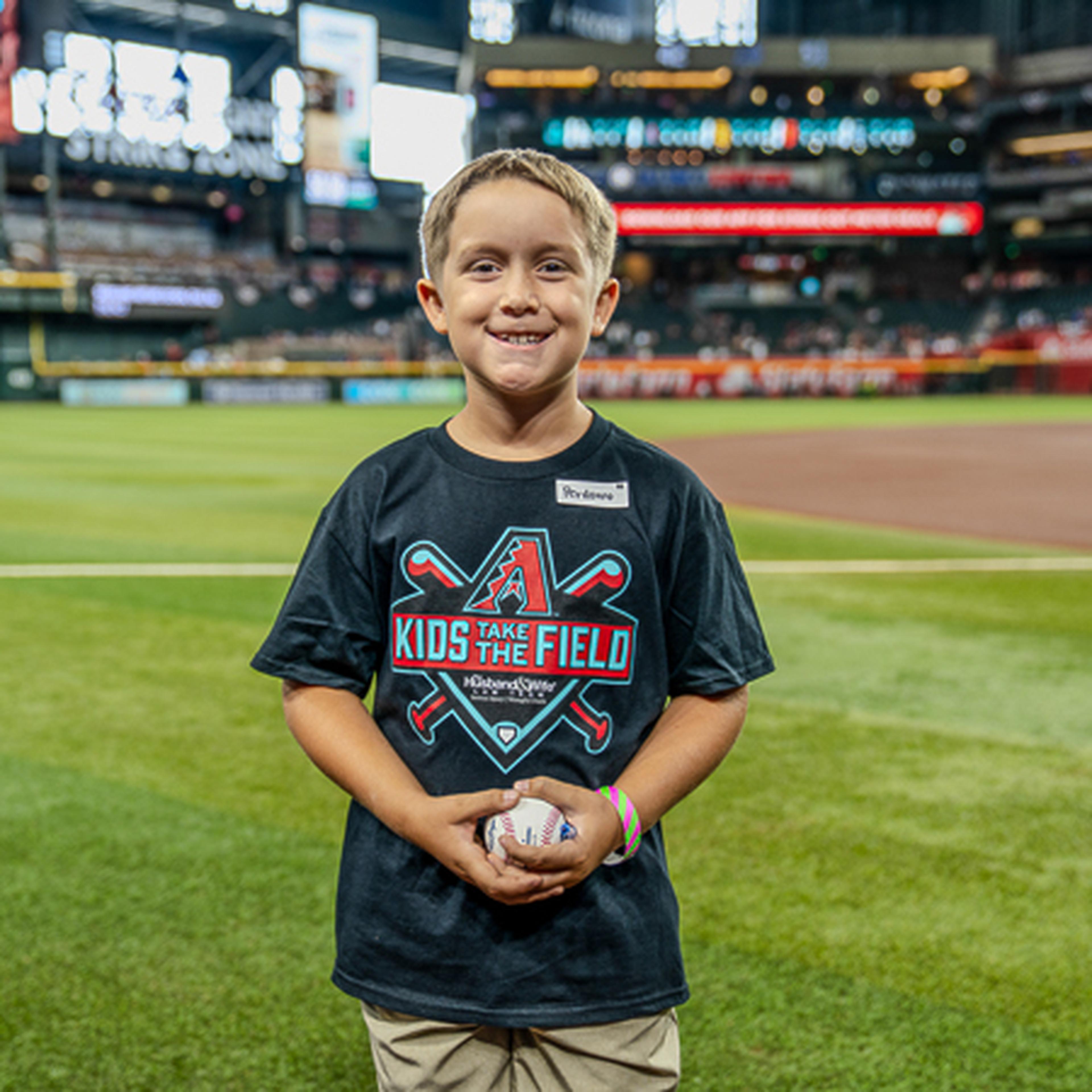 Smiling boy stands on a baseball field wearing a "Kids Take the Field" T-shirt, holding a baseball. Stadium seats in the background.