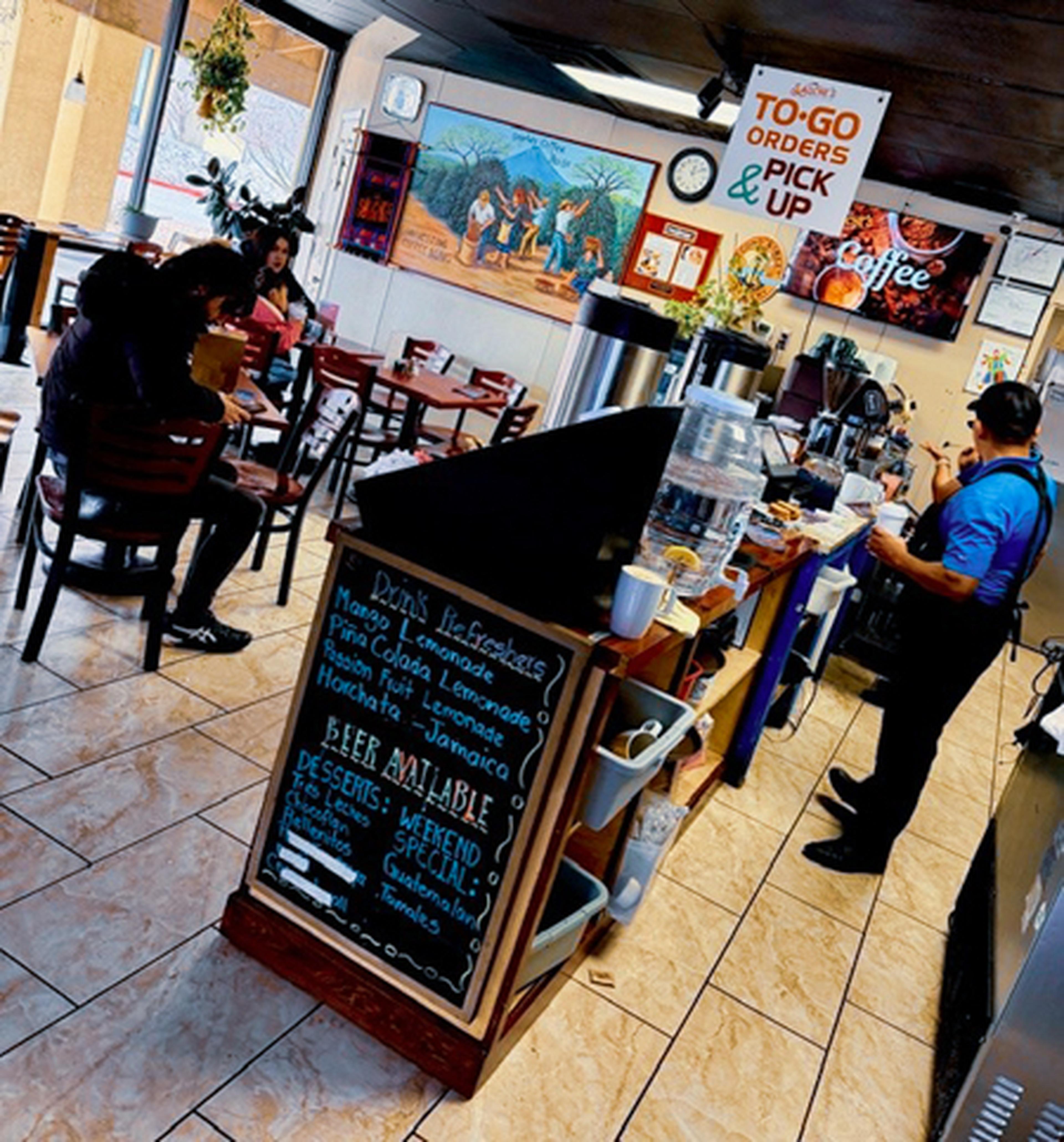 Inside a small café, people sit at tables eating. A chalkboard menu lists beverages. A barista prepares drinks behind the counter.