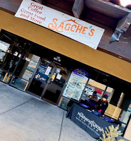 Entrance of Sagche's Coffee House with Lisa stands behind a table covered with Husband & Wife Law Team materials