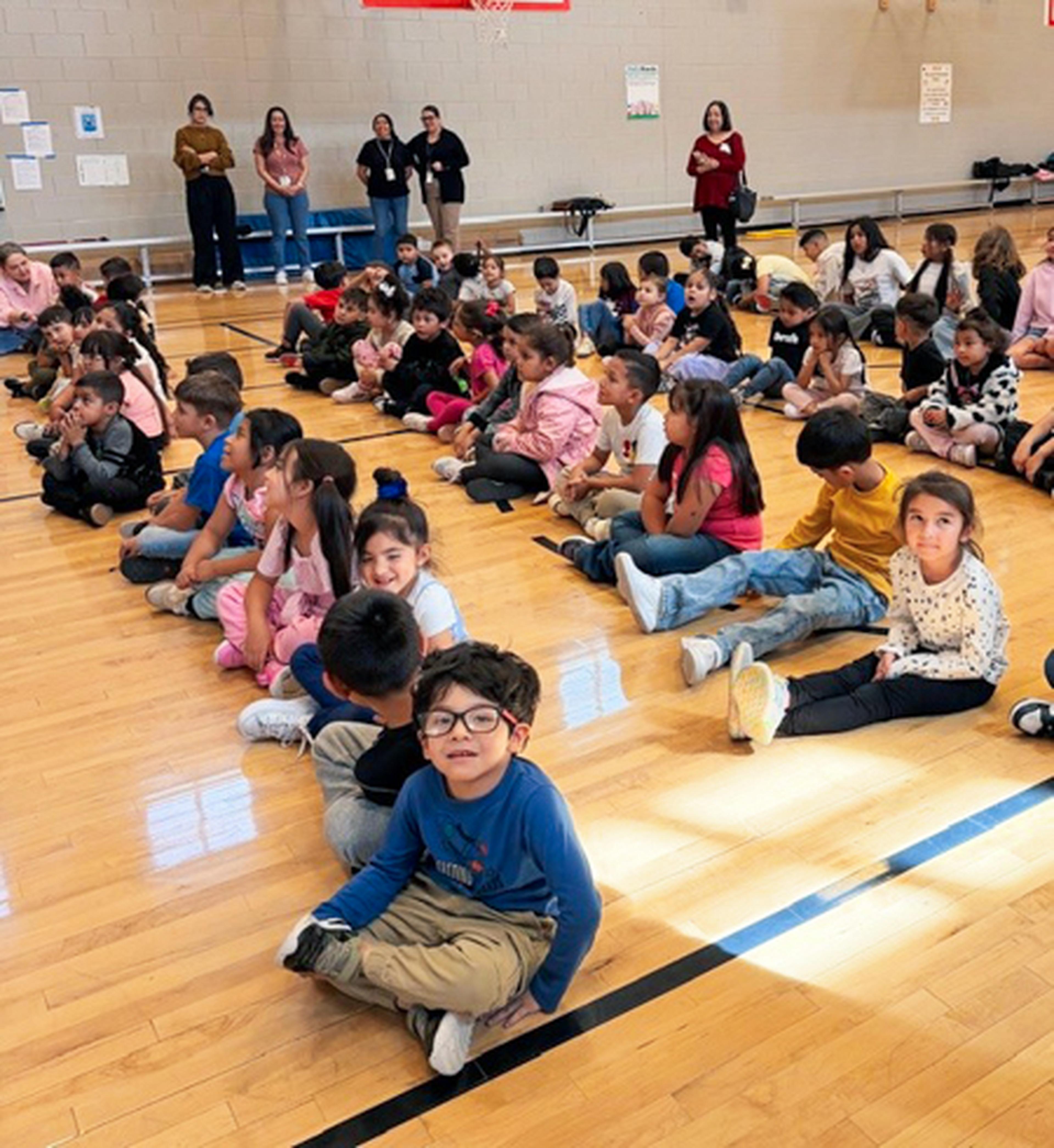 Third-grade kids sitting on a gym floor in neat rows, with adults nearby