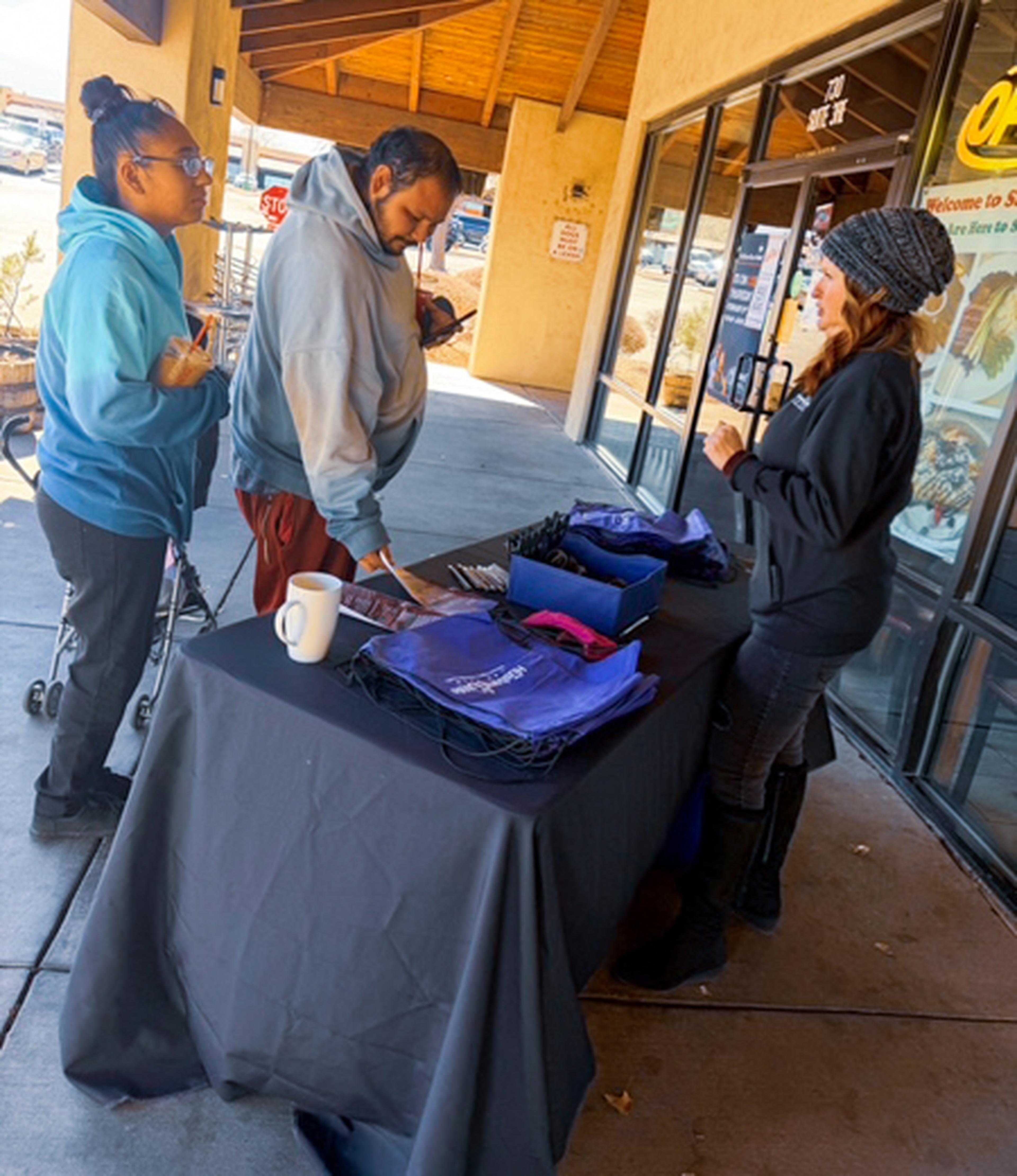 Two people engage with a woman at an outdoor table featuring bags, papers, and a mug, under a wooden awning next to a building entrance.