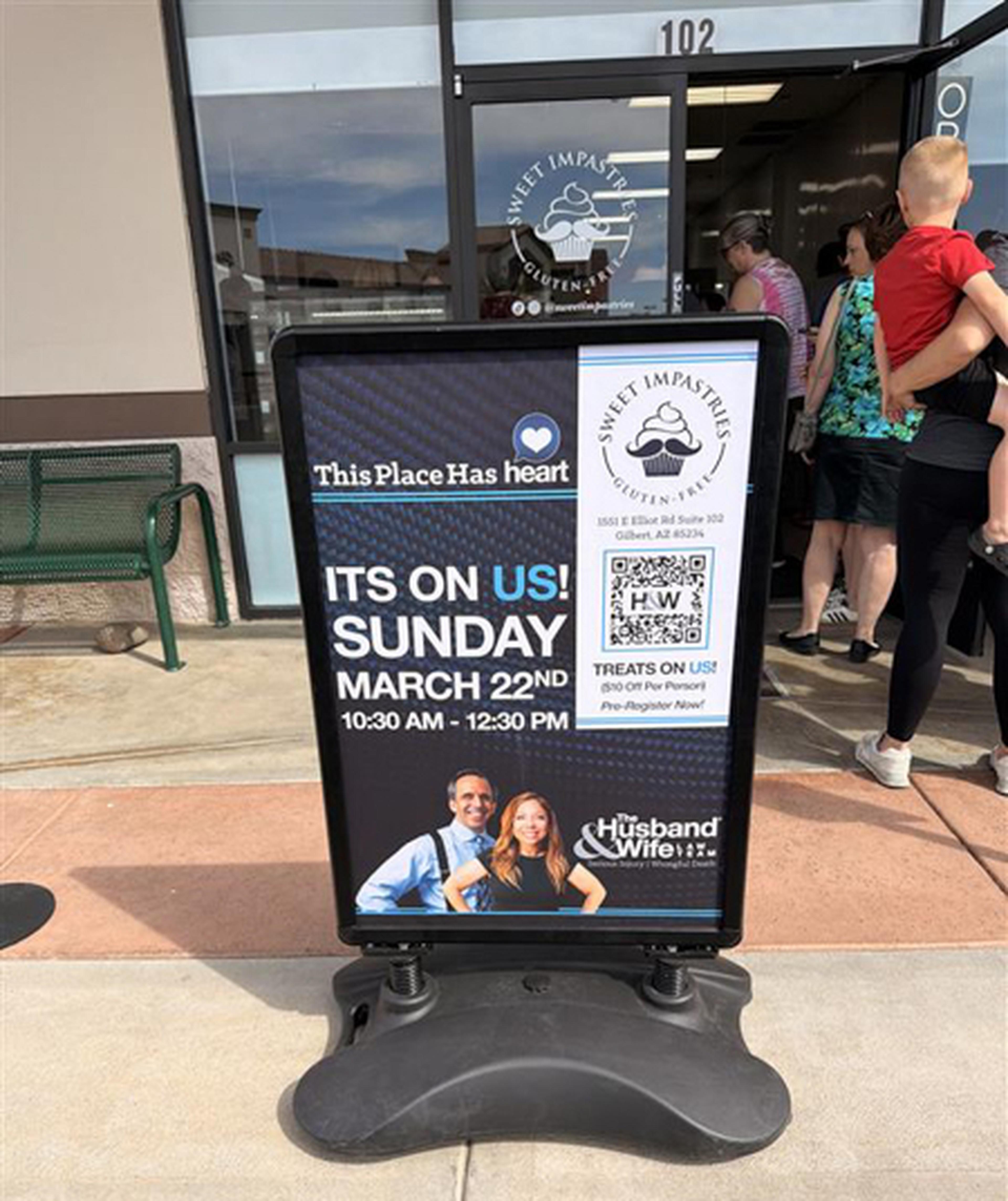 A Husband & Wife Law Team sign stands outside Sweet Impastries, promoting a free March 22nd event with treats and a QR code, as customers line up at the bakery.