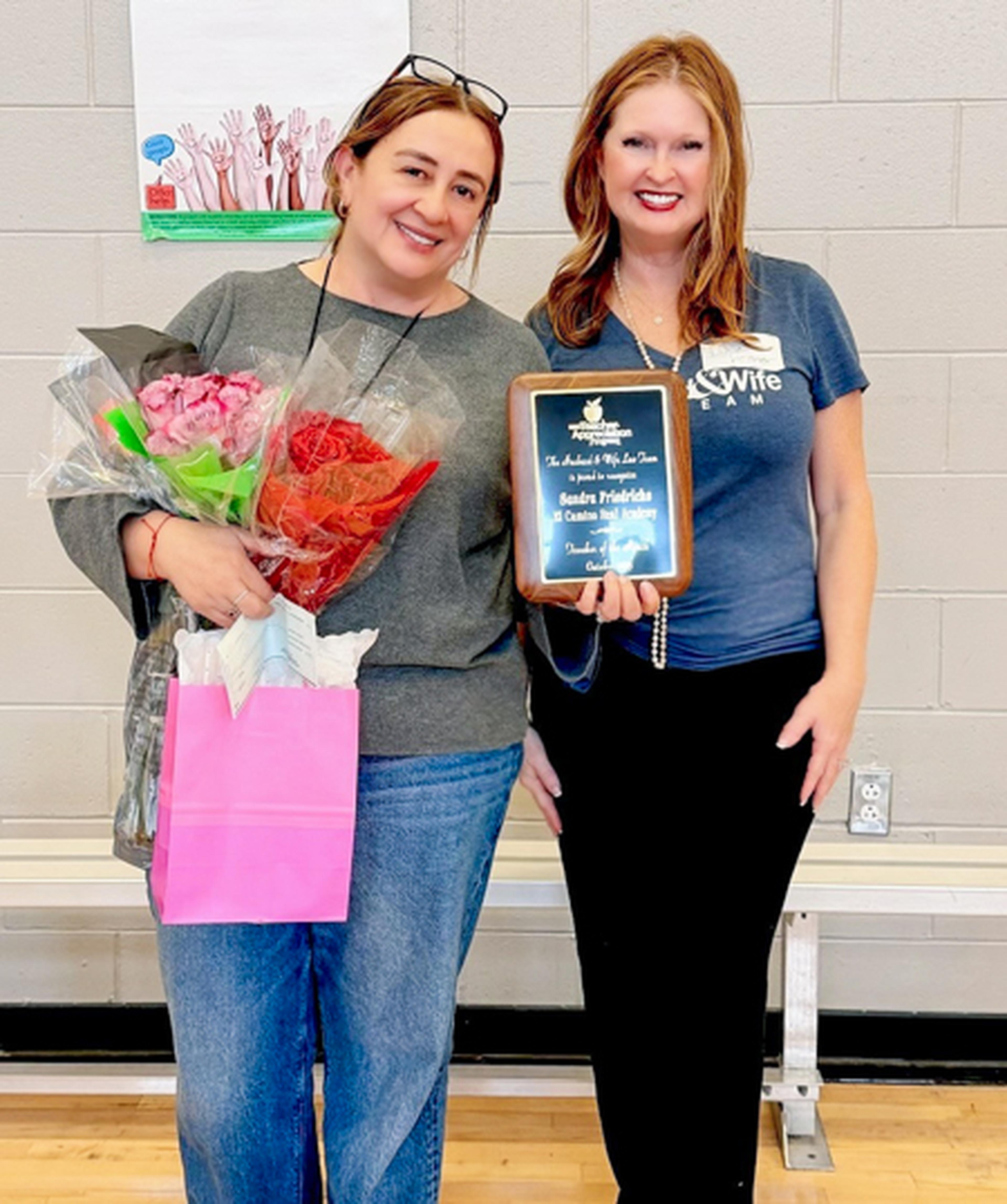 Two women pose for a photo, one is holding a plaque and the other is holding two bunches of roses and a pink bag