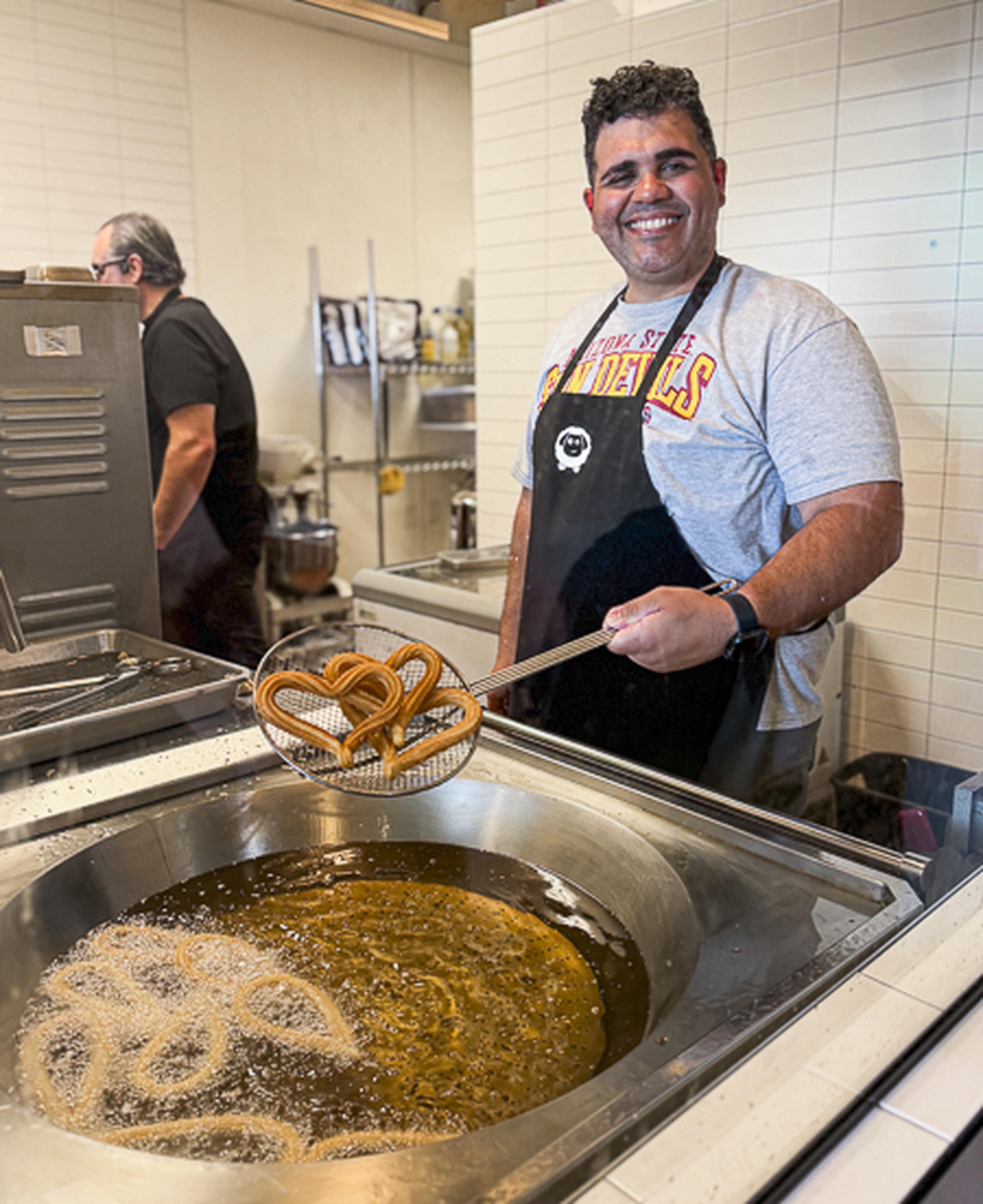 A grinning churro‑maker lifts a strainer of fresh, golden churros like treasure, ready to delight whoever’s waiting.
