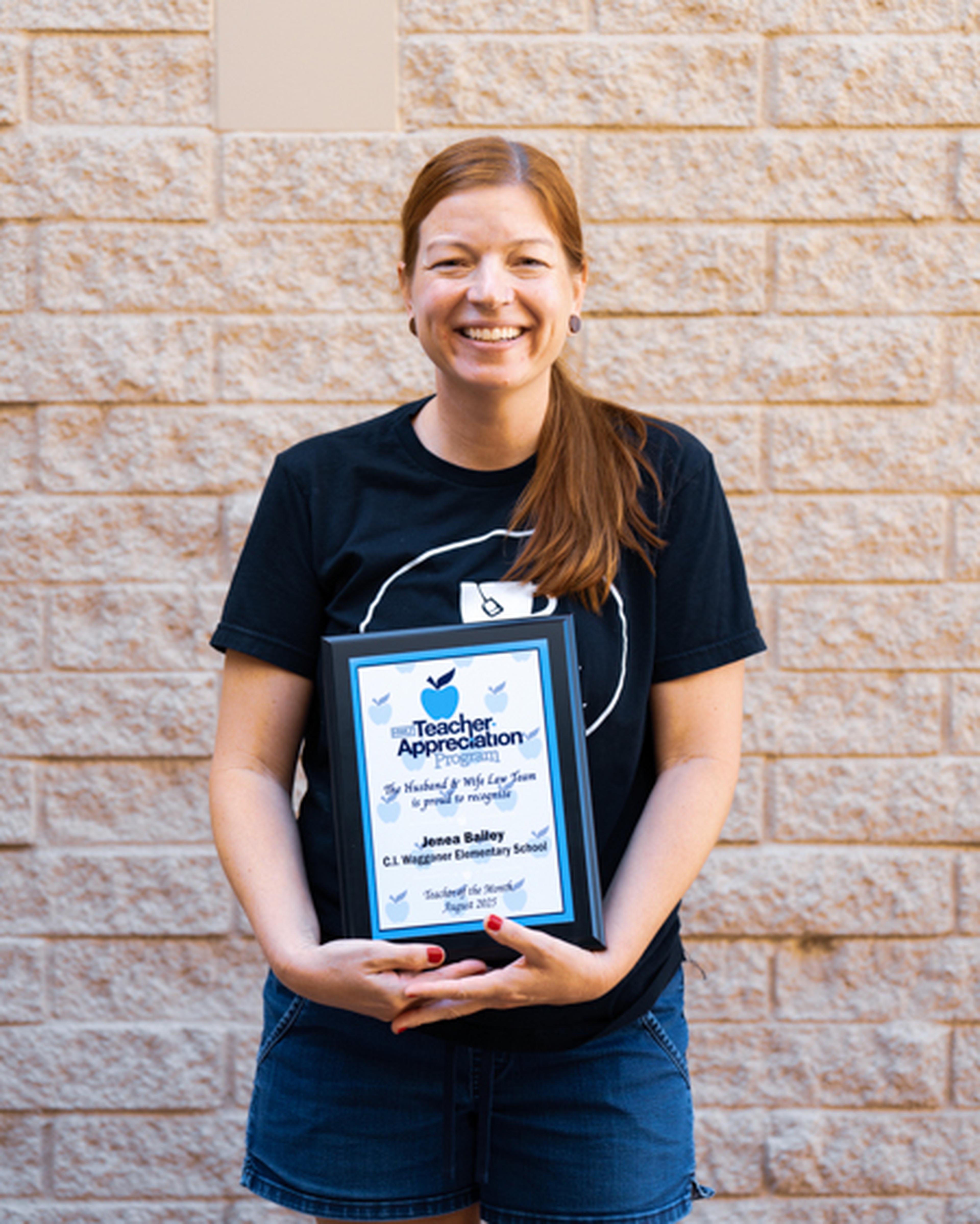 Mrs. Bailey smiles as she stands outside the school with her Teacher Appreciation plaque