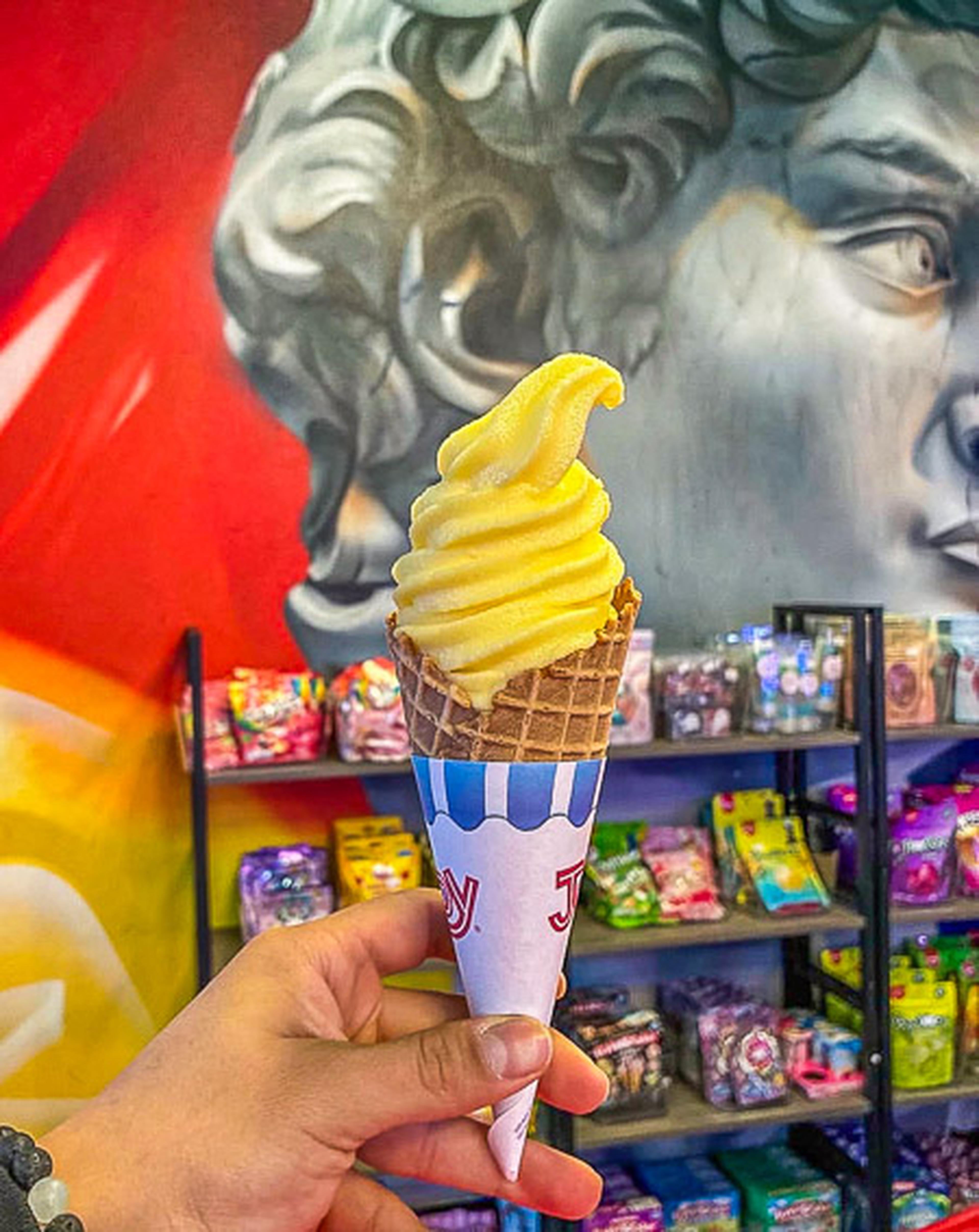 Hand holding a yellow soft-serve ice cream cone in front of a colorful mural and shelves of candy in a shop.