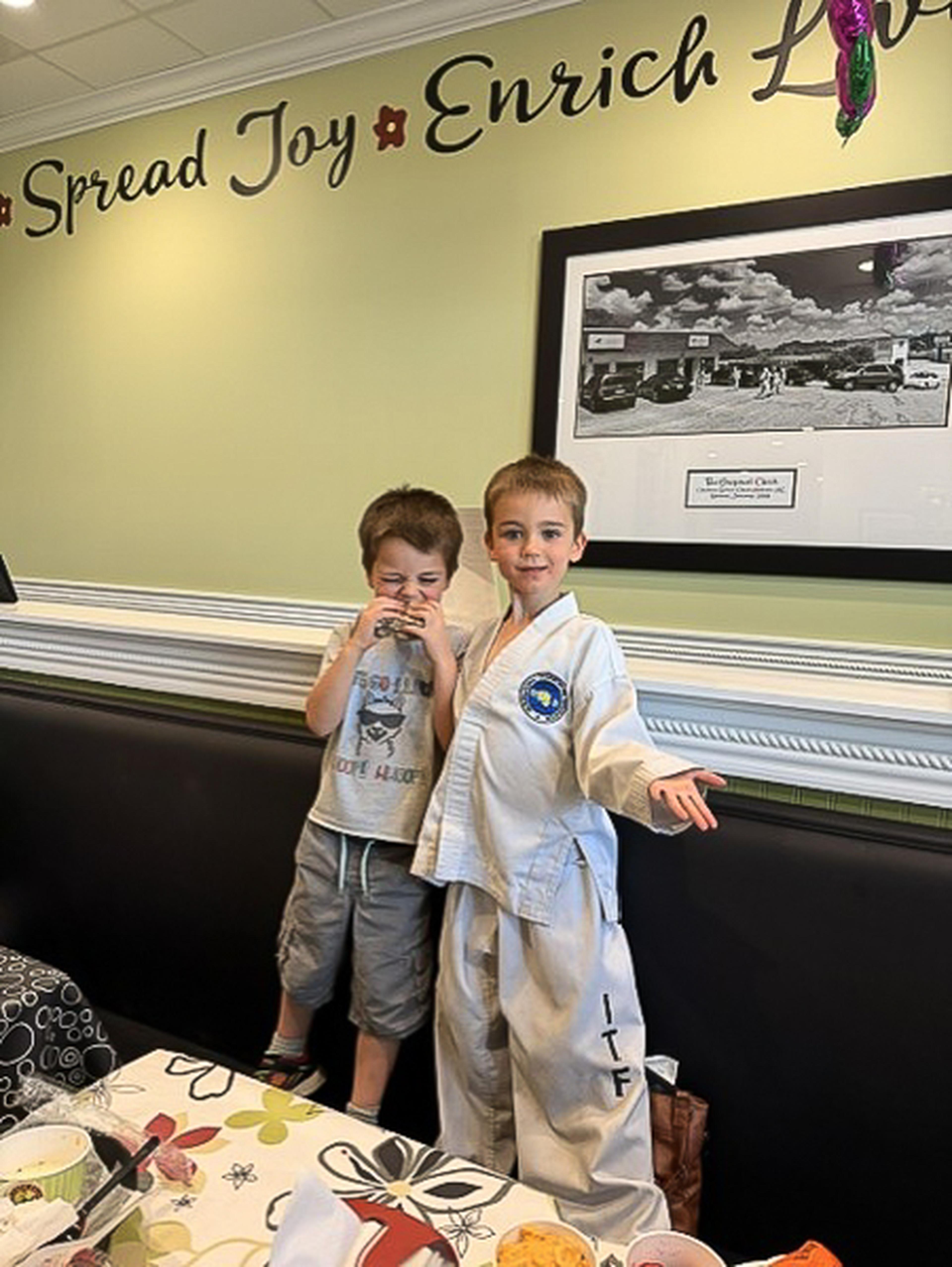 Two young boys stand on a booth seat inside Chicken Salad Chick, one dressed in a martial arts uniform, beneath the wall message “Spread Joy Enrich Love.”