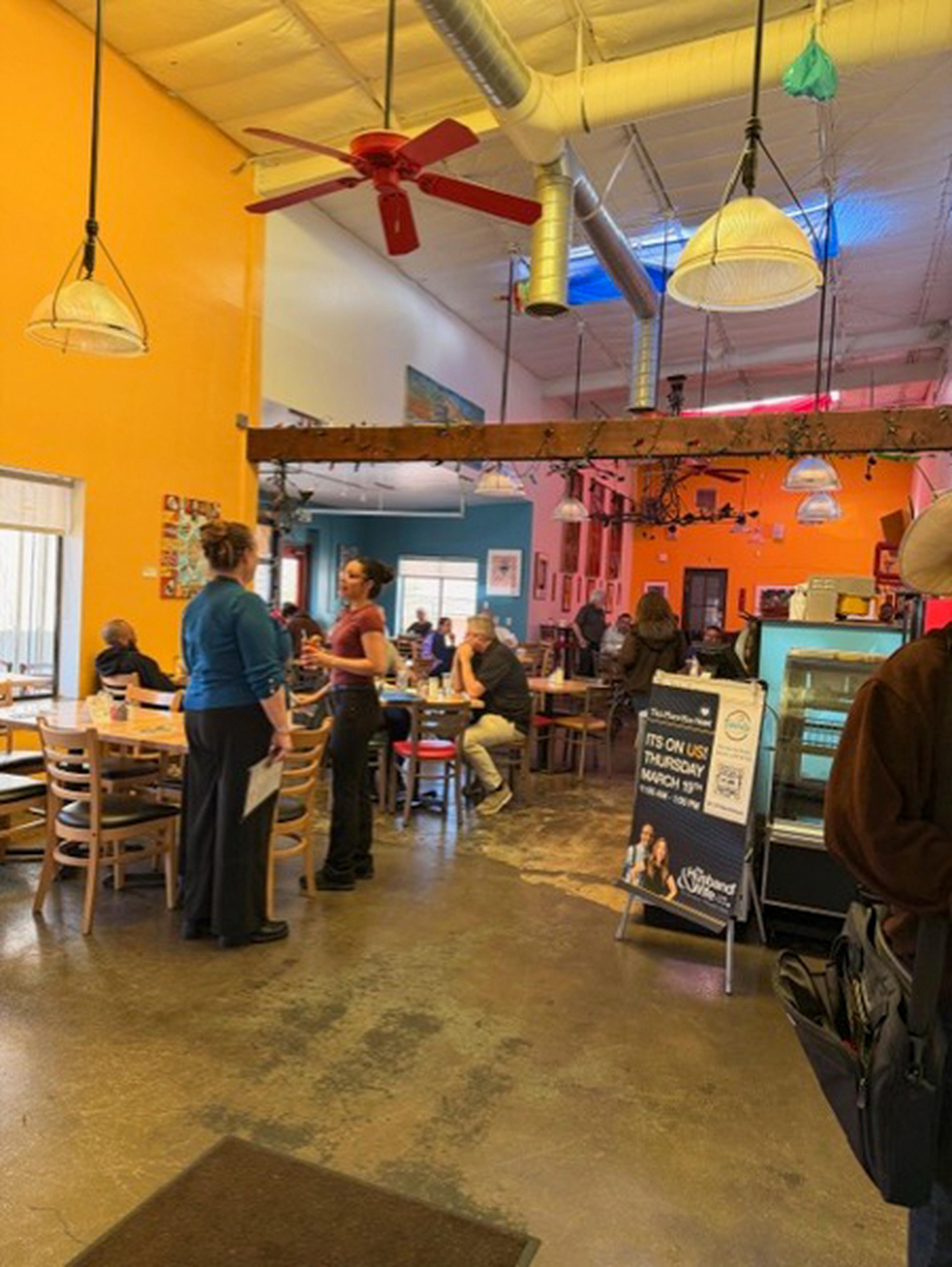 The Bonita Restaurant is a vibrant cafe with orange walls, patrons seated at wooden tables, and staff interacting. Ceiling fans and hanging lights add to the decor.