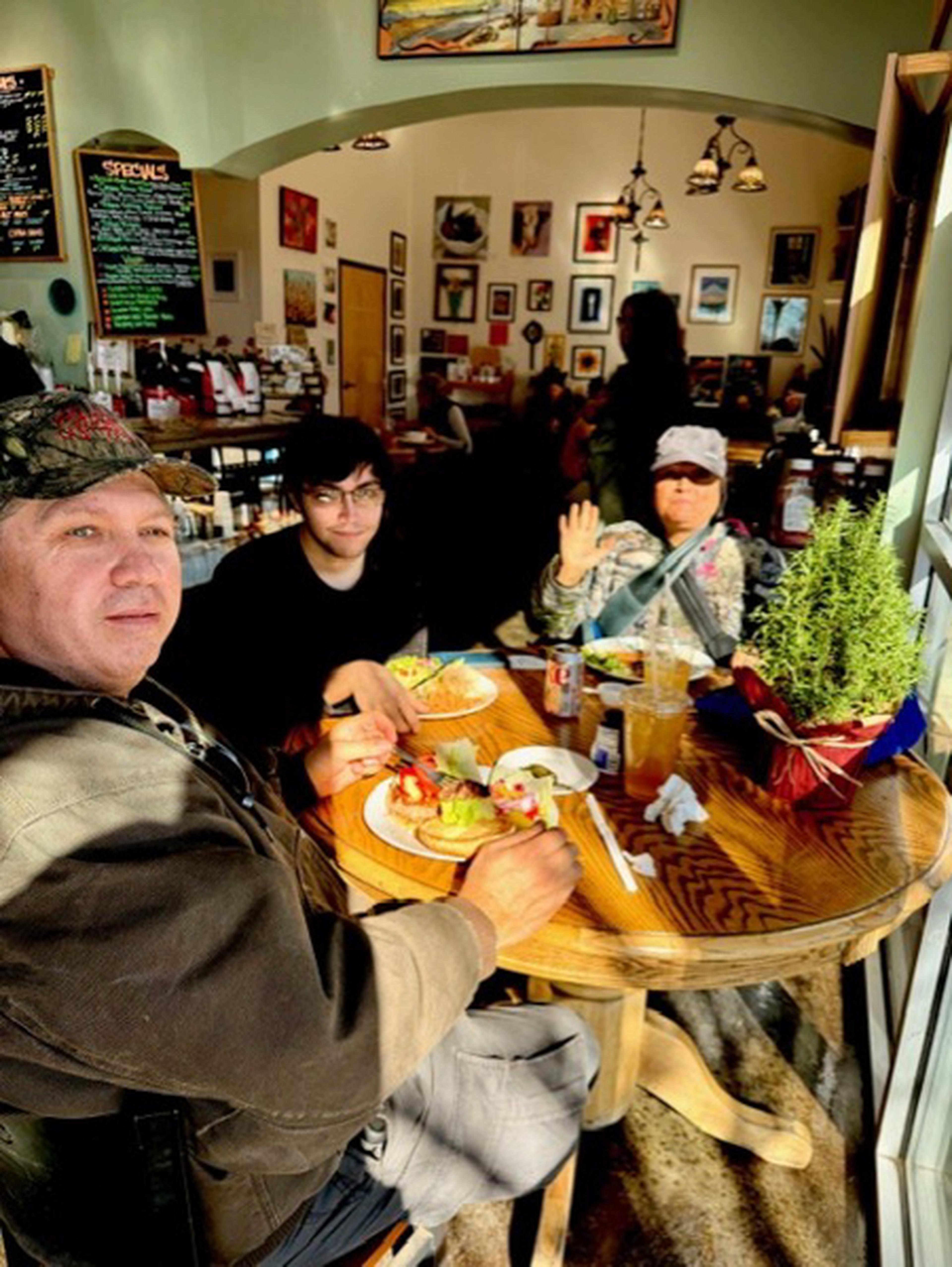 Three people sitting at a wooden table in at Tribes Coffeehouse, eating a meal. The room is decorated with framed pictures and a menu board on the wall.