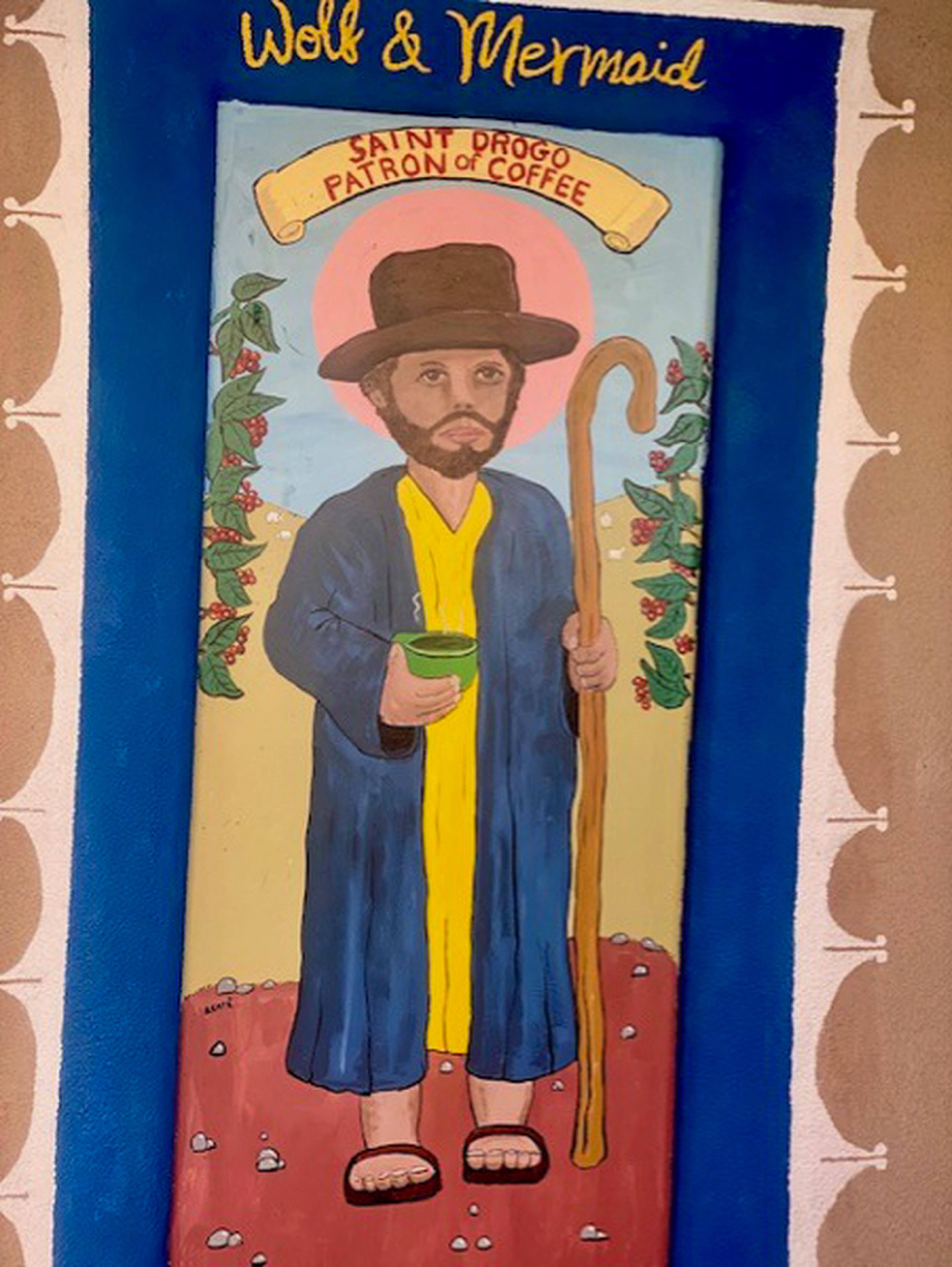 Painting of Saint Drogo holding a staff and green cup, labeled "Patron of Coffee," with birds and greenery in the background.