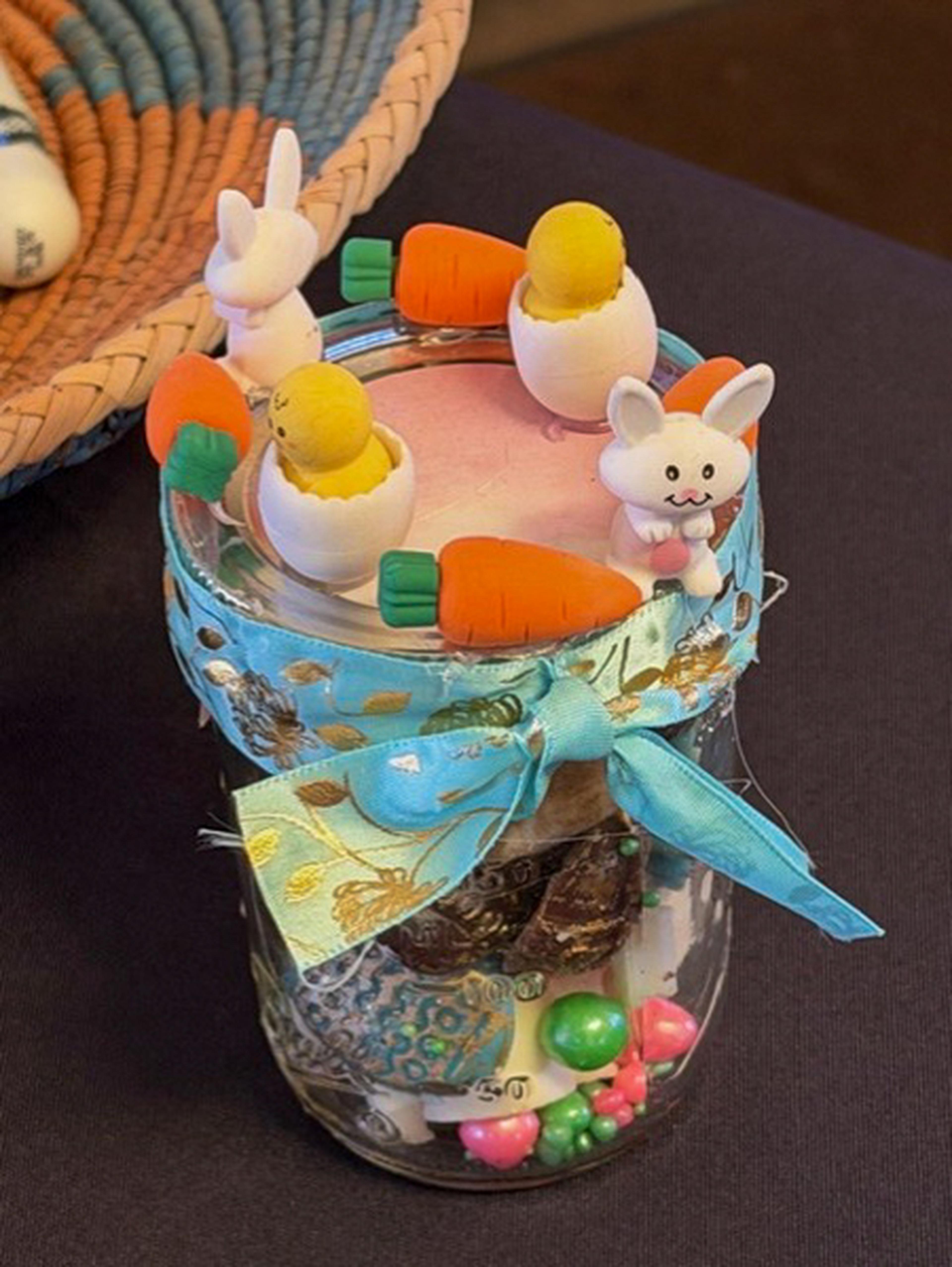 A decorated jar with a teal bow, bunny and carrot figures, and colorful marbles inside, set on a dark surface.
