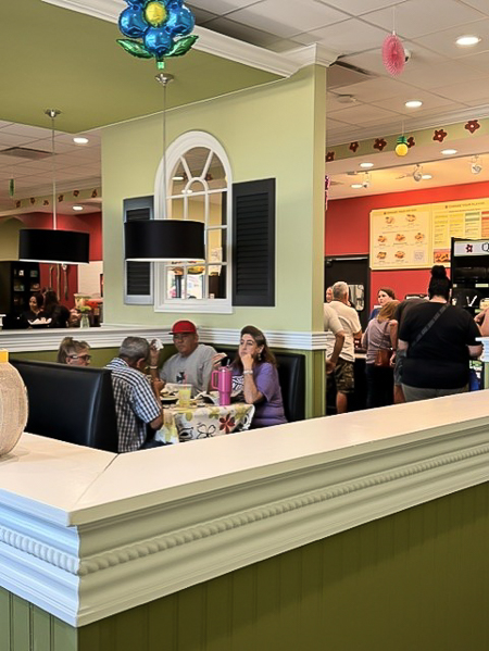 Customers dine and chat inside Chicken Salad Chick, a cozy café filled with colorful décor and hanging lights, with a group gathered at the counter in the background.