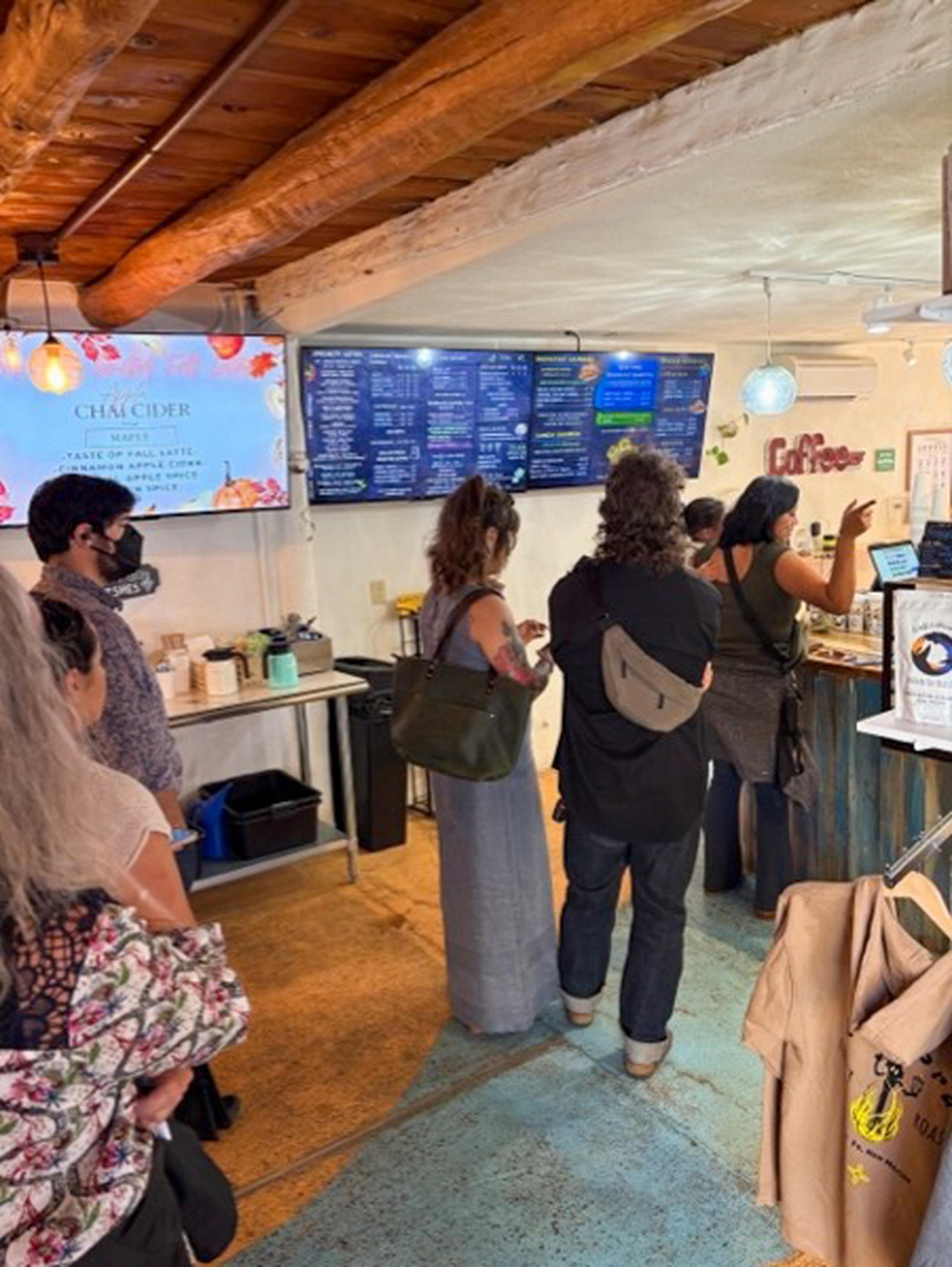 People standing in line at a cozy café with wooden beams, viewing a digital menu above the counter displaying various coffee and beverage options.