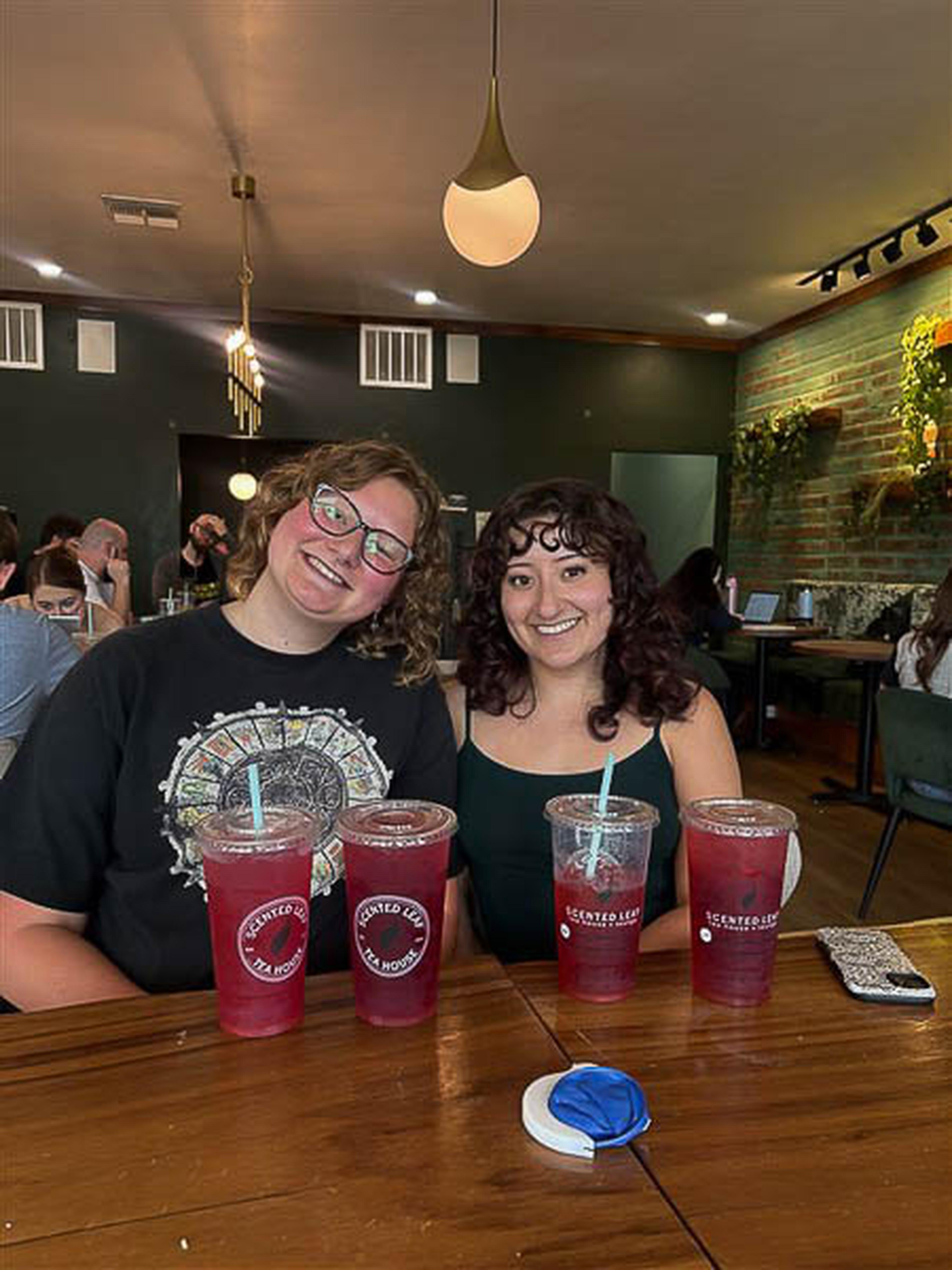 Two friends smile at Scented Leaf in Tucson, iced drinks in hand and greenery filling the lively space.