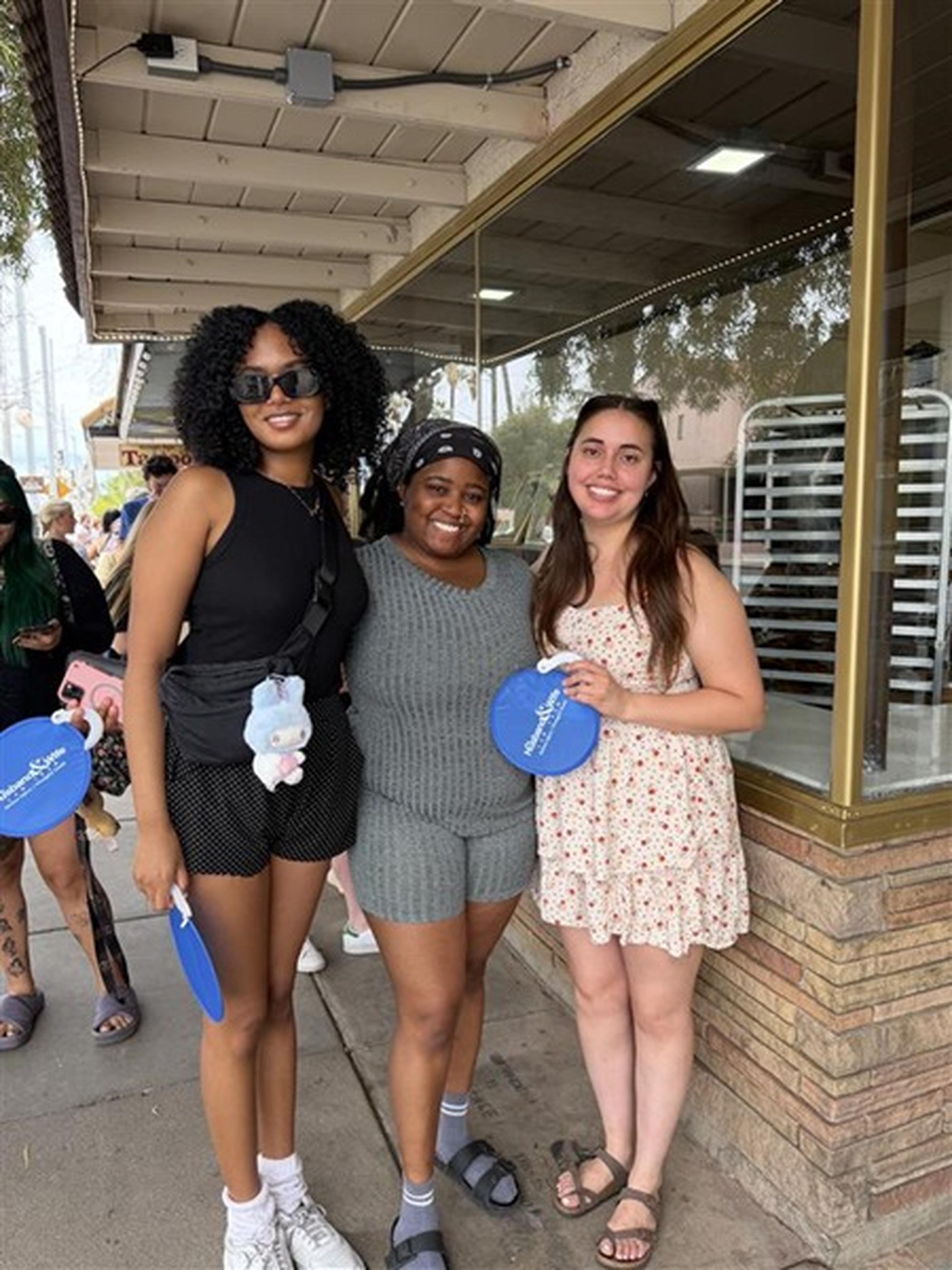 Three friends smile on the sidewalk, holding blue Husband & Wife Law Team fans, with a brick building and others behind them.