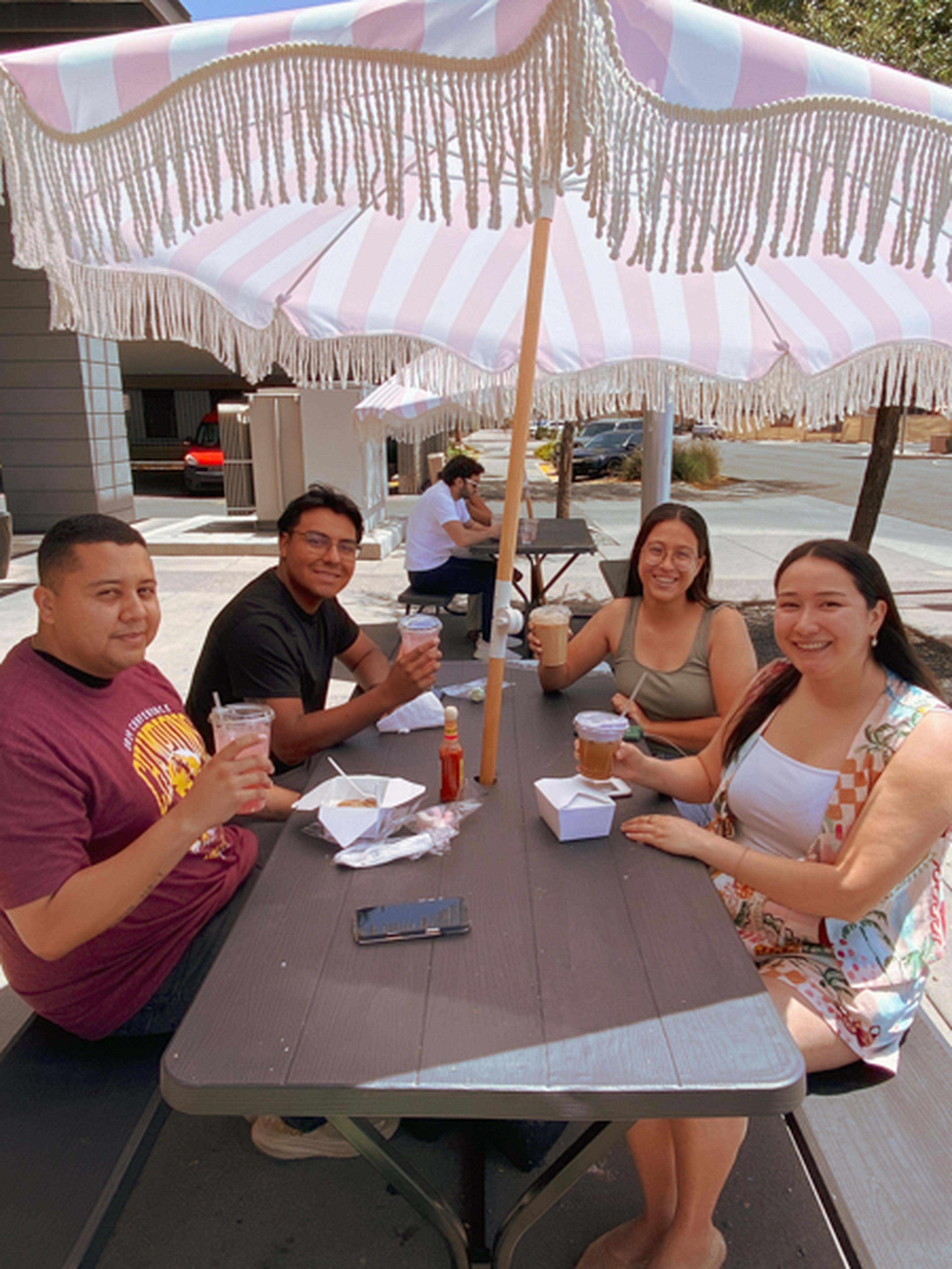 Four people sitting under a pink and white umbrella outdoors with their brunches.