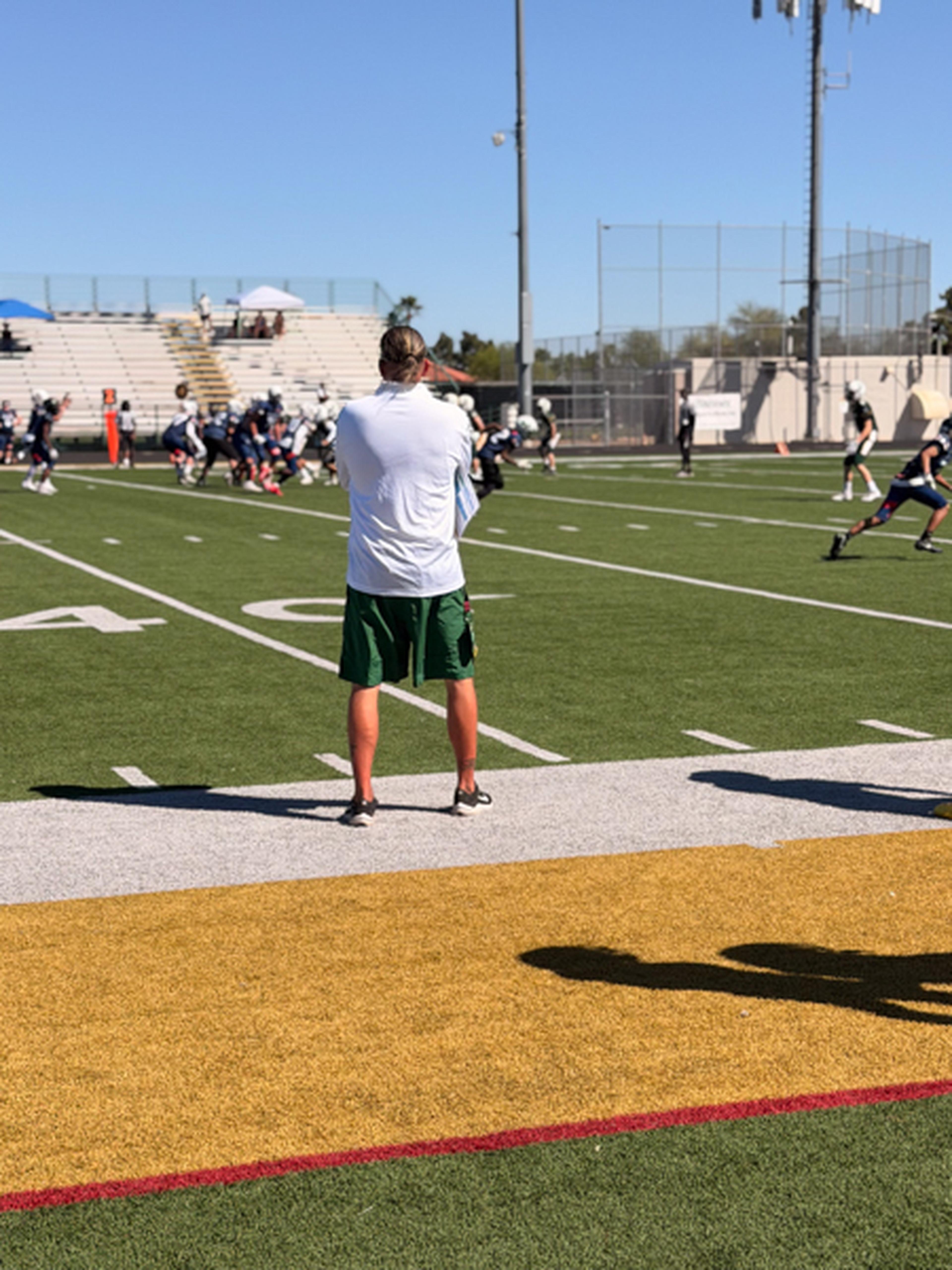 Coach Tomas, in a white shirt and green shorts, watches the game from the sideline on a sunny day with players and bleachers in view.