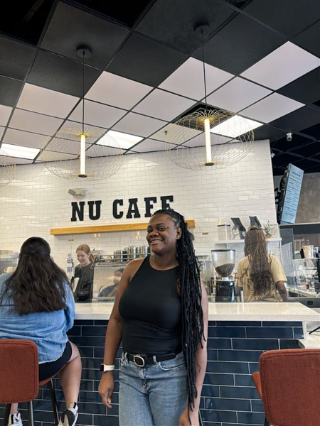 The owner of NU Cafe smiles as the event is a huge success! Woman in a black top and jeans stands smiling in a cafe with a white tiled wall and "NU CAFE" sign in the background.