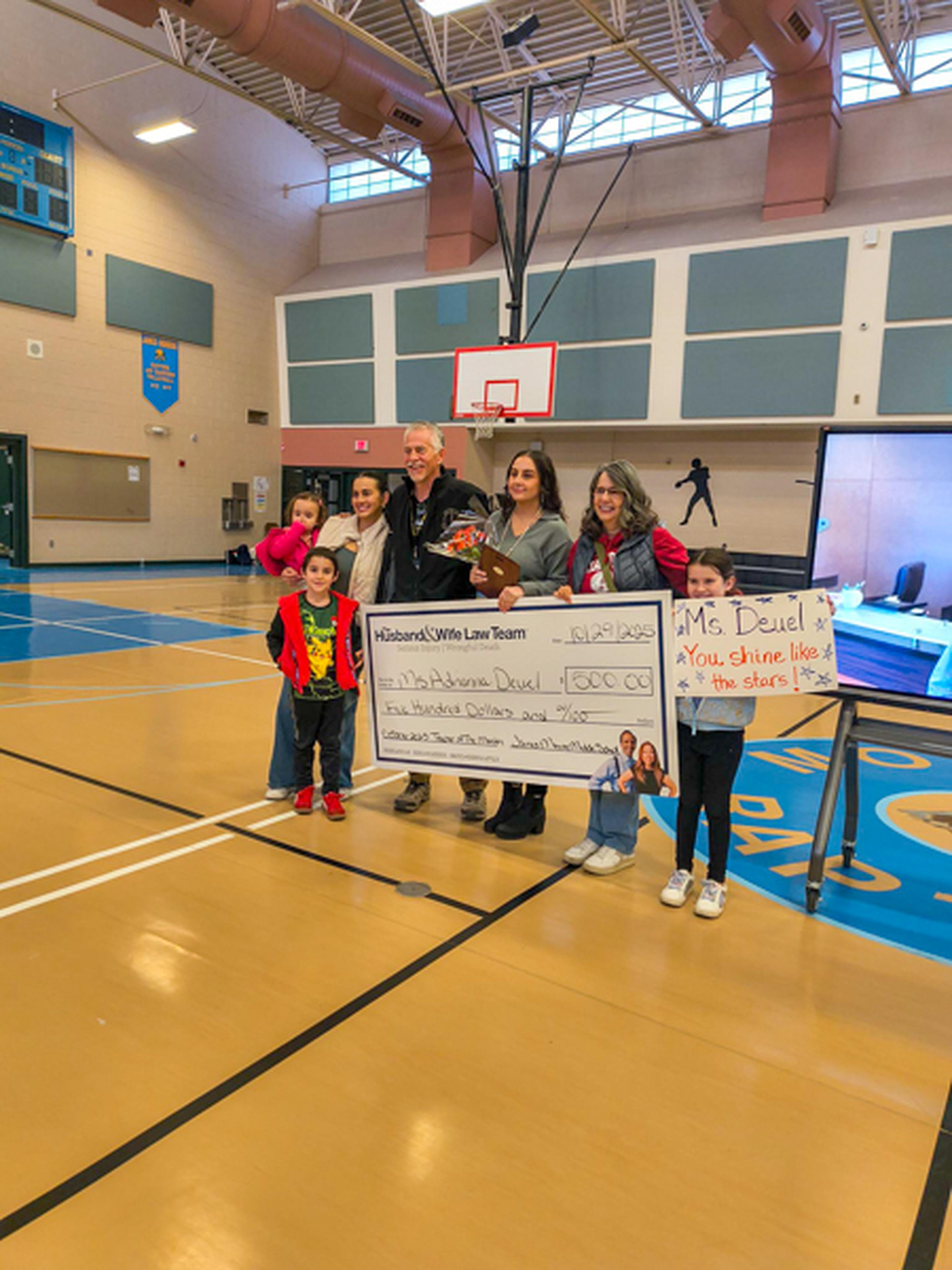 People holding a large check and sign in a gymnasium, celebrating with smiles. Basketball hoop and scoreboard visible in the background.