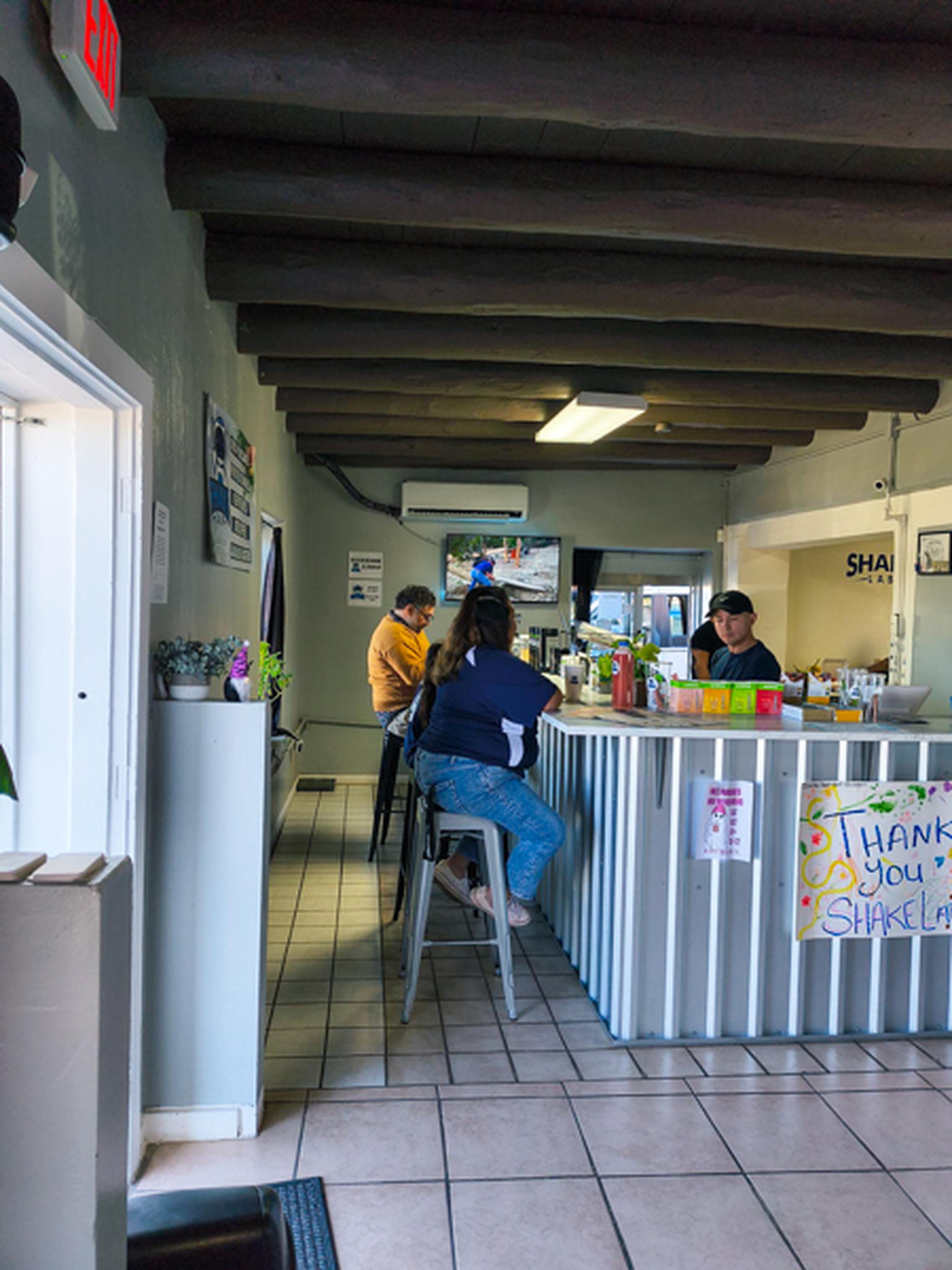 People sitting at a counter in a small cafe with wooden beams, tiled floor, and a TV on the wall. Bright, welcoming atmosphere.