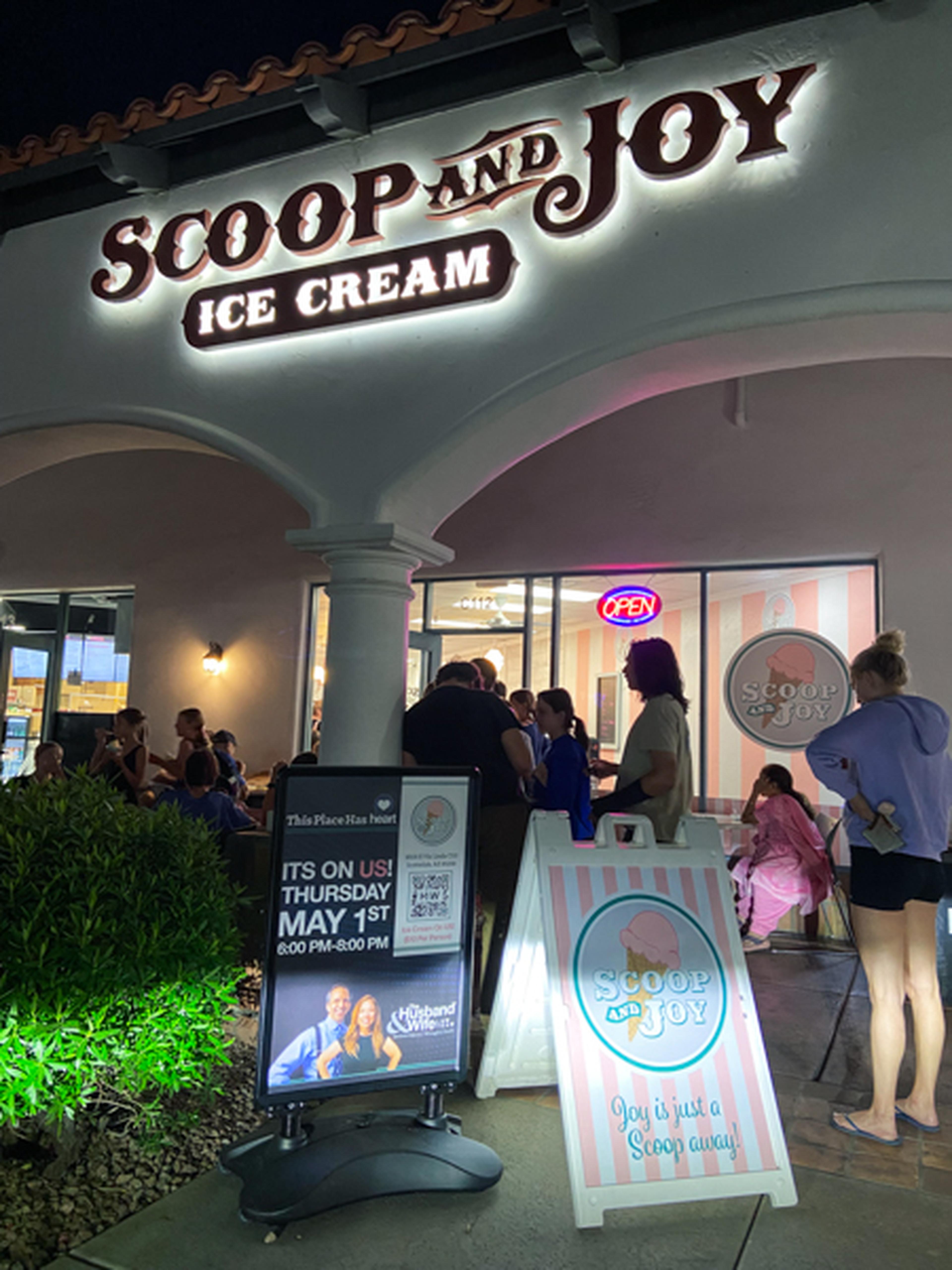 People gather outside "Scoop and Joy Ice Cream" shop at night, with a sign announcing a special event with The Husband & Wife Law Team