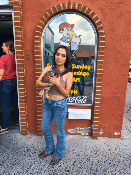 Do they have tacos for ducks? Woman standing in front of a brick-arched restaurant window, holding a duck, cartoon chef logo visible on the glass.