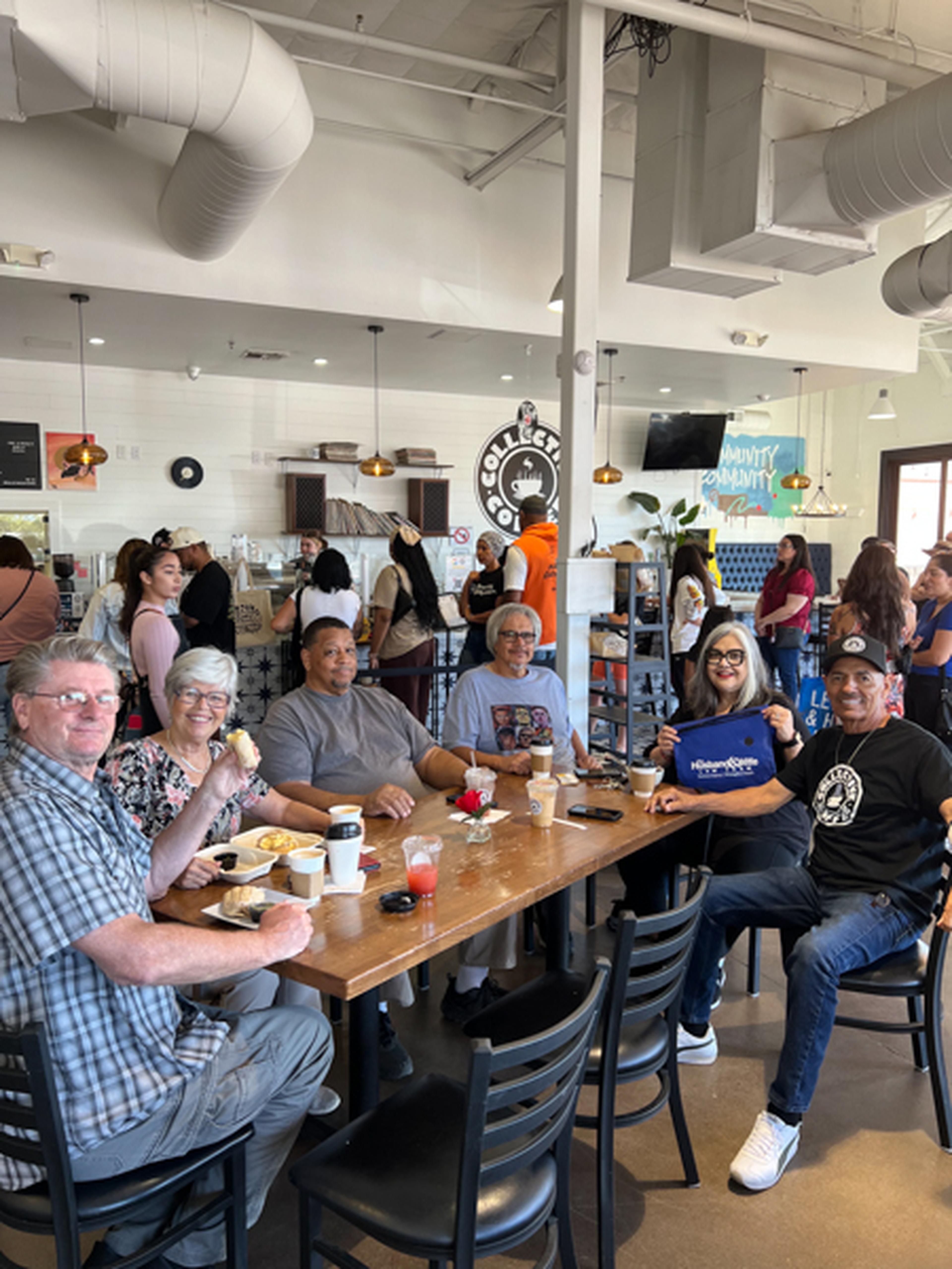 A group of people smiling and sitting around a table in a busy cafe, enjoying food and drinks.