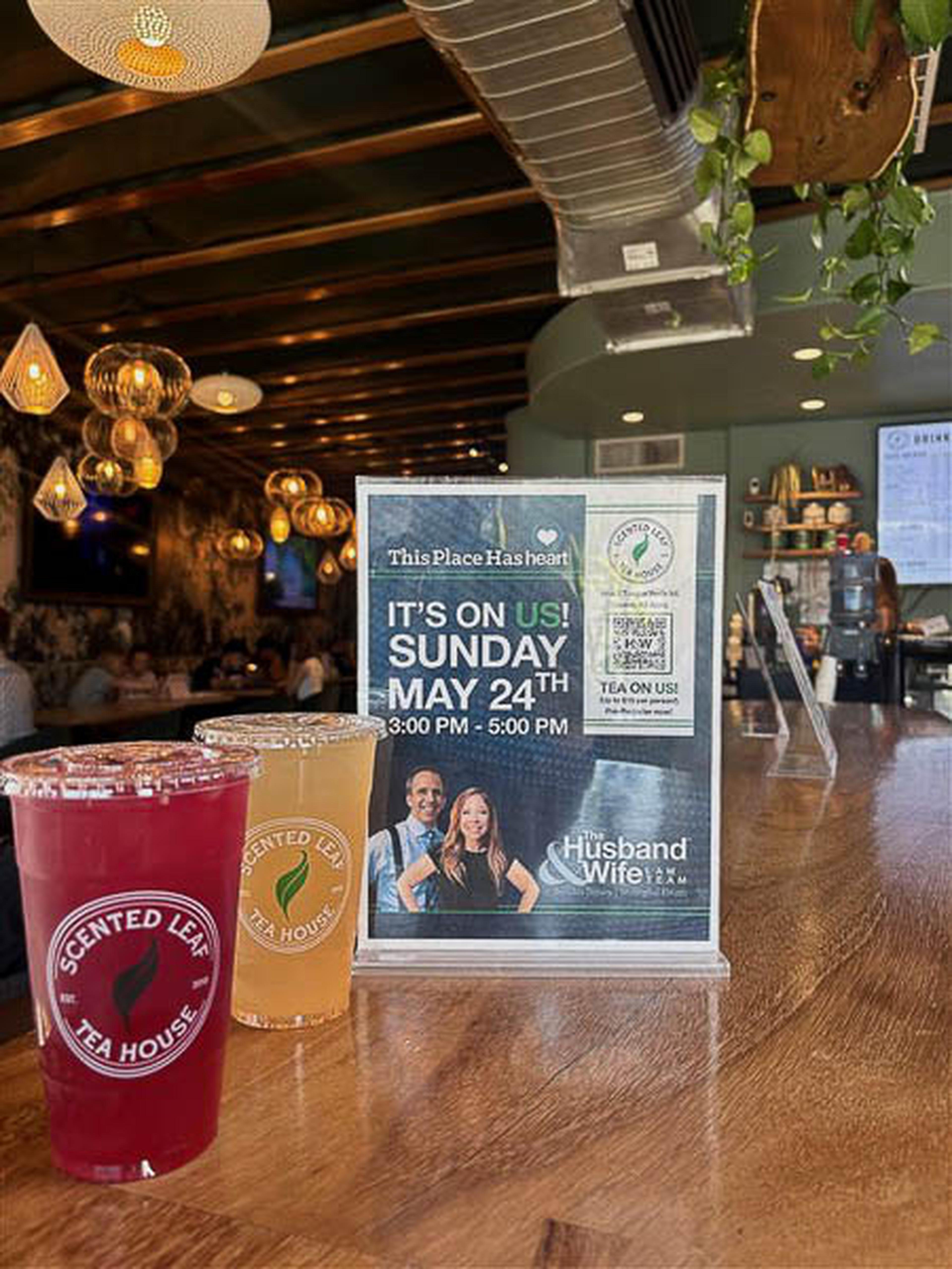 Two iced teas rest on a wooden counter beside a sign for a May 24th tea event hosted by the Husband & Wife Law Team, glowing under cozy hanging lights.