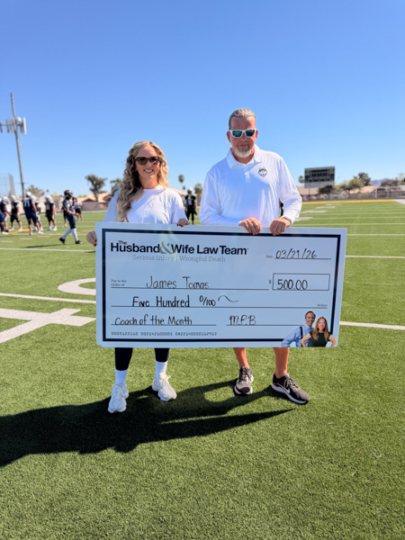 Jen from the Husband & Wife Law Team and Coach Tomas hold a $500 “Coach of the Month” check awarded to James Tomas on the football field.