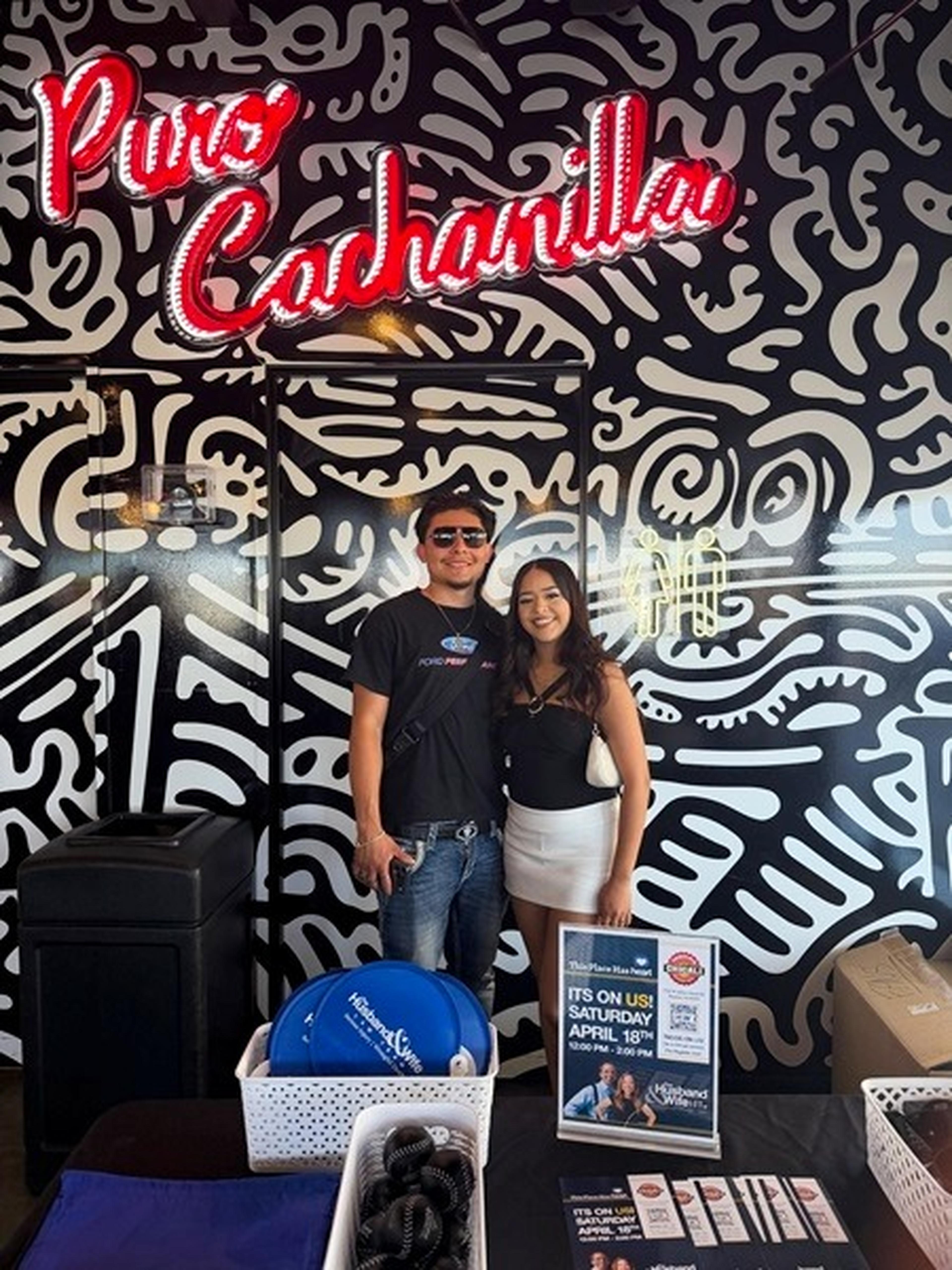 Two people stand smiling in front of a decorative black-and-white mural under a red neon sign reading "Puro Cachanilla", with promotional items on a table.