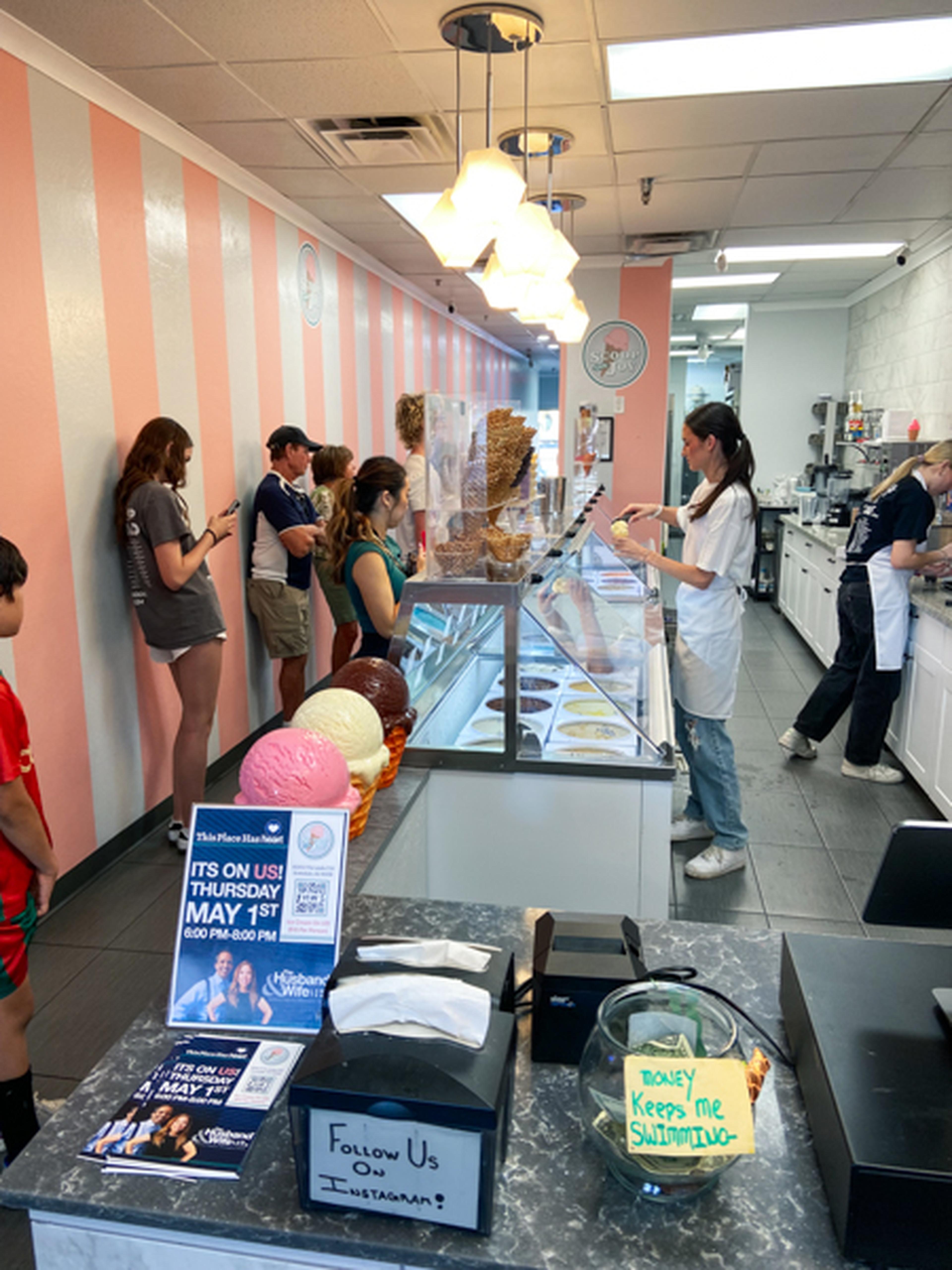 Customers line up inside an ice cream shop with pink-striped walls, choosing flavors from a glass display. Staff serve behind the counter.