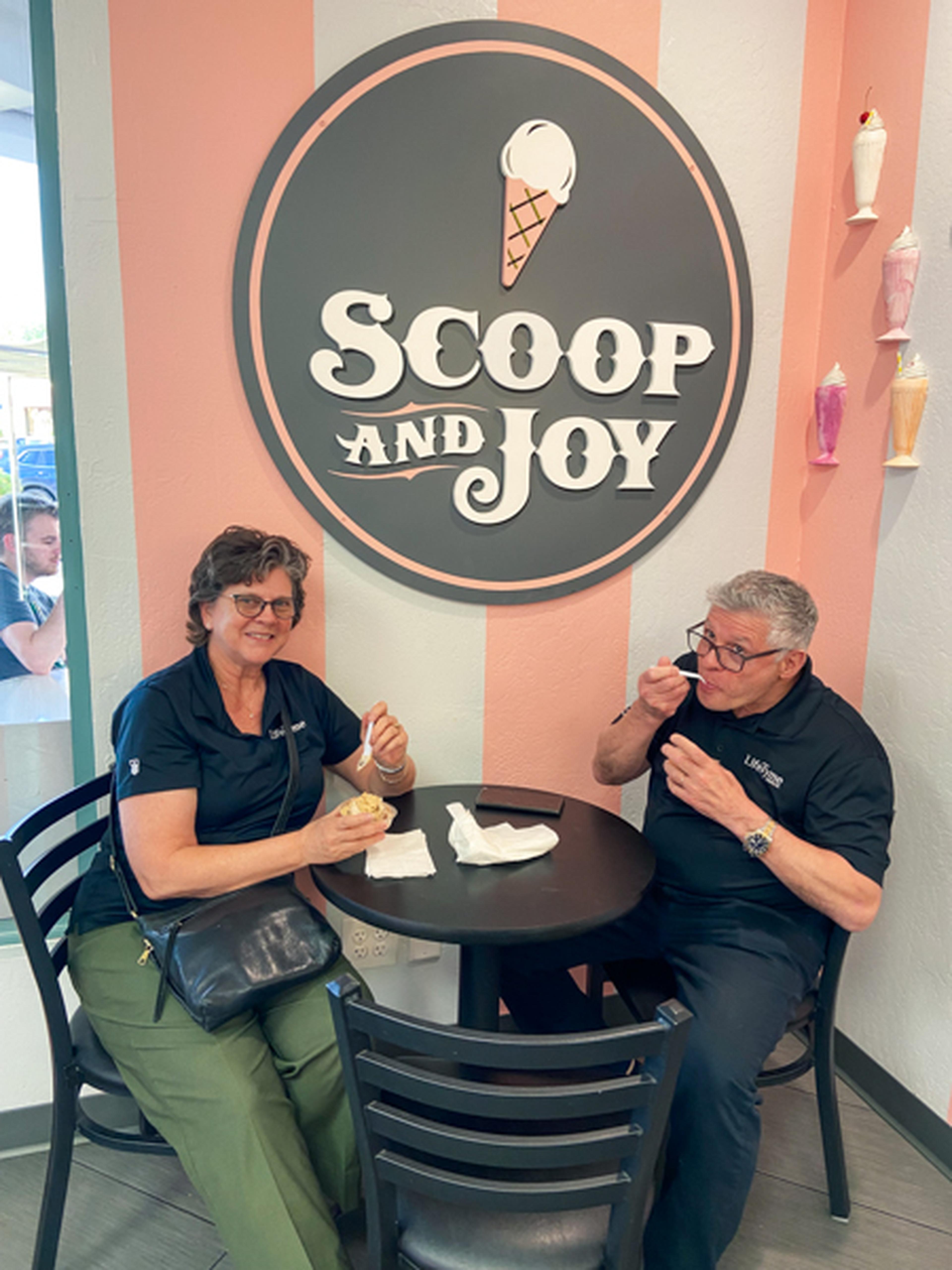 Two people enjoying ice cream at a table in an ice cream shop. A sign on the wall reads "Scoop and Joy" with ice cream decorations.
