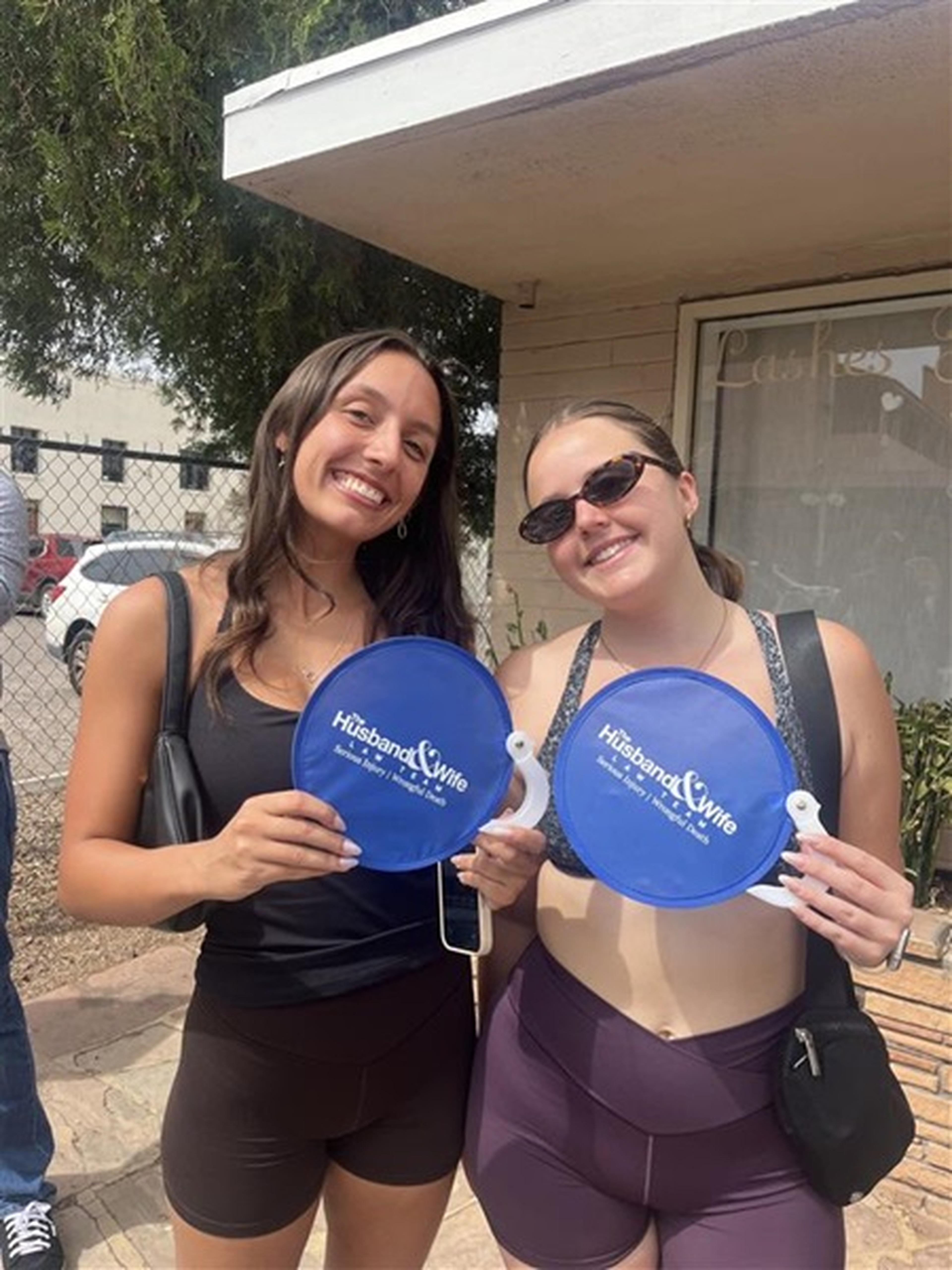 Two smiling women outdoors hold blue fans labeled "Husband & Wife," wearing casual summer attire and sunglasses.