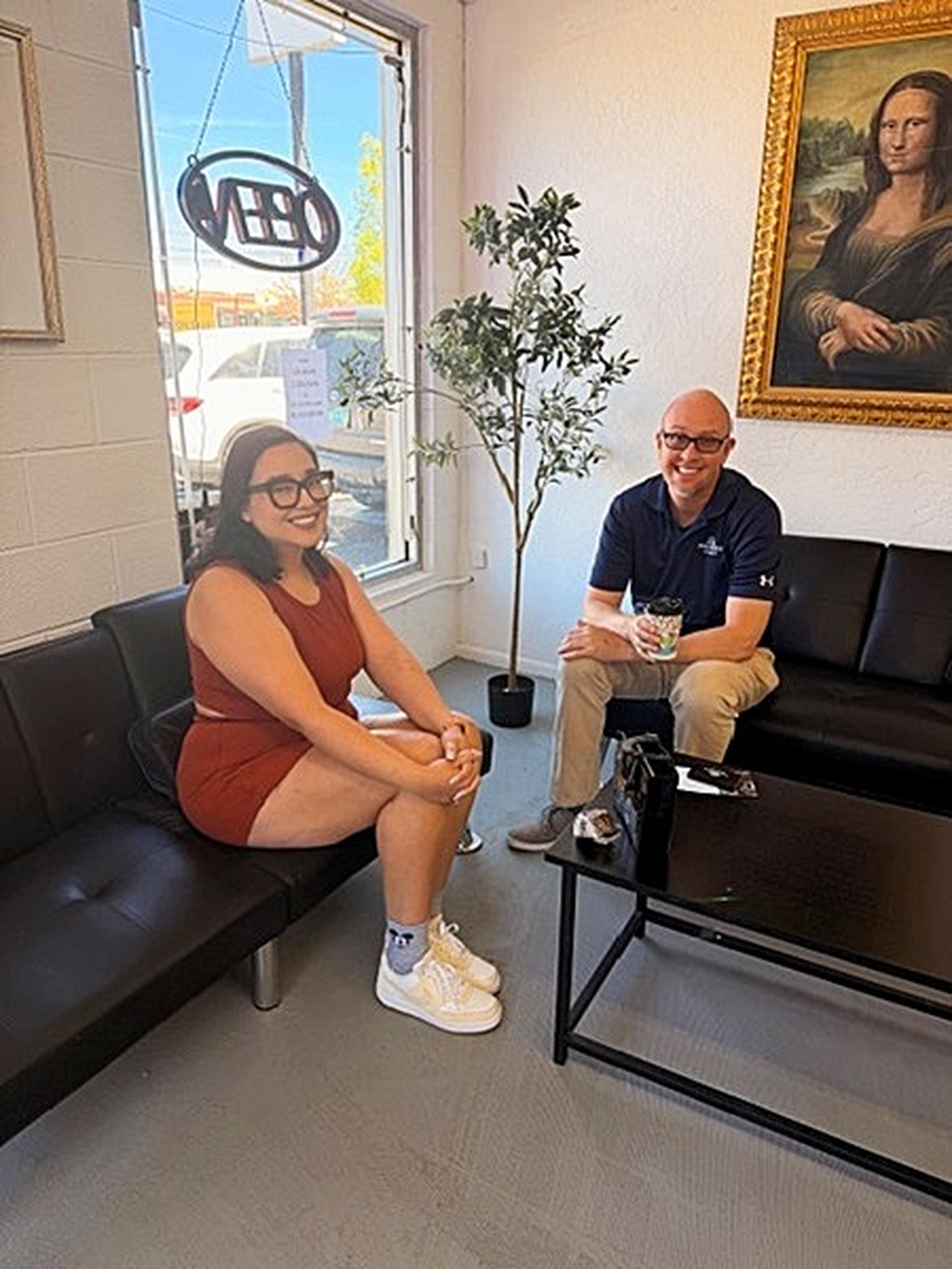 Two people smile while relaxing on black sofas at Milk and Honey Caffe, framed by a potted plant and a Mona Lisa painting.