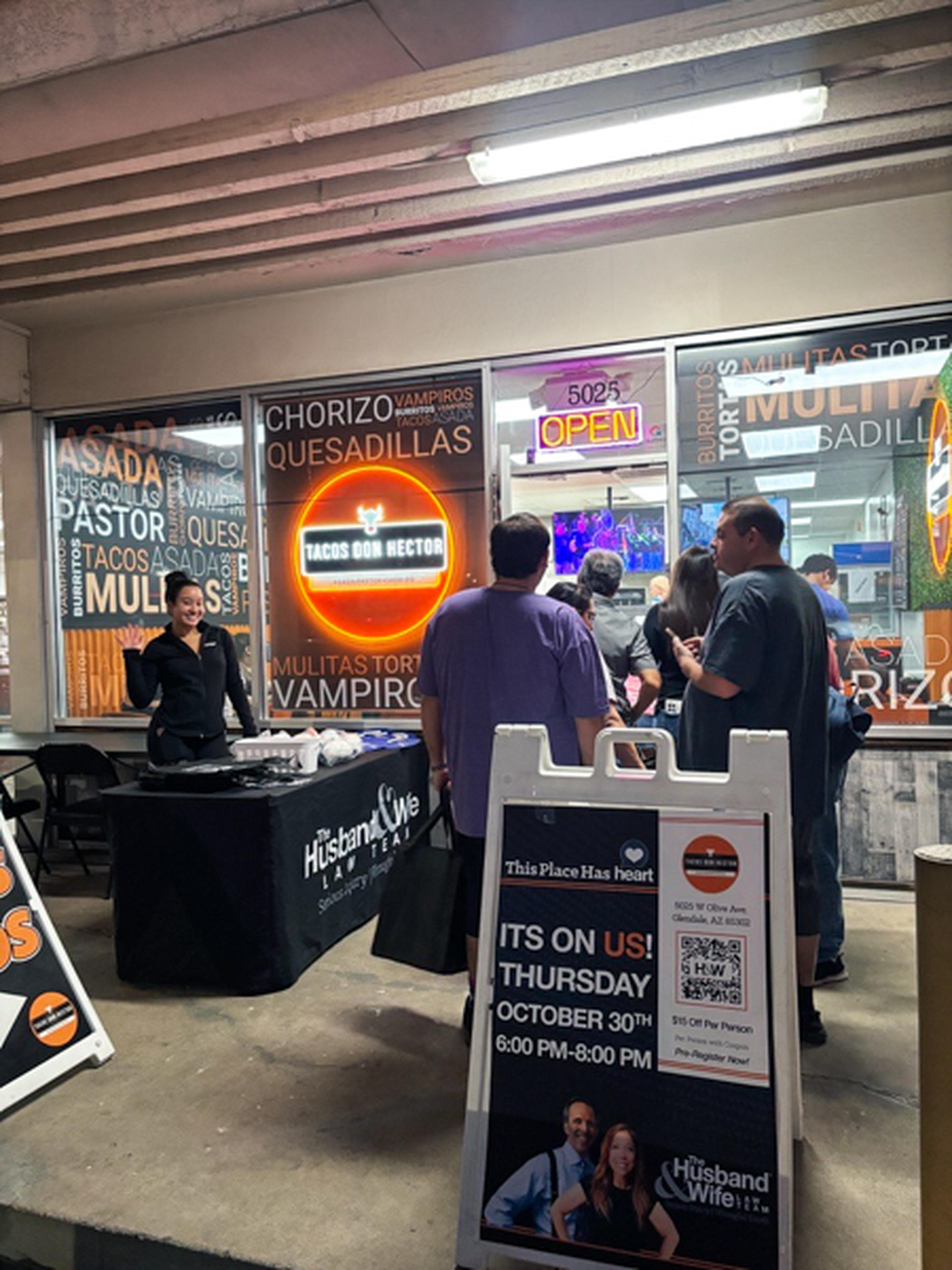 People gathered outside a taco shop with neon signs, a promotional stand, and a signboard for an event on October 30th.