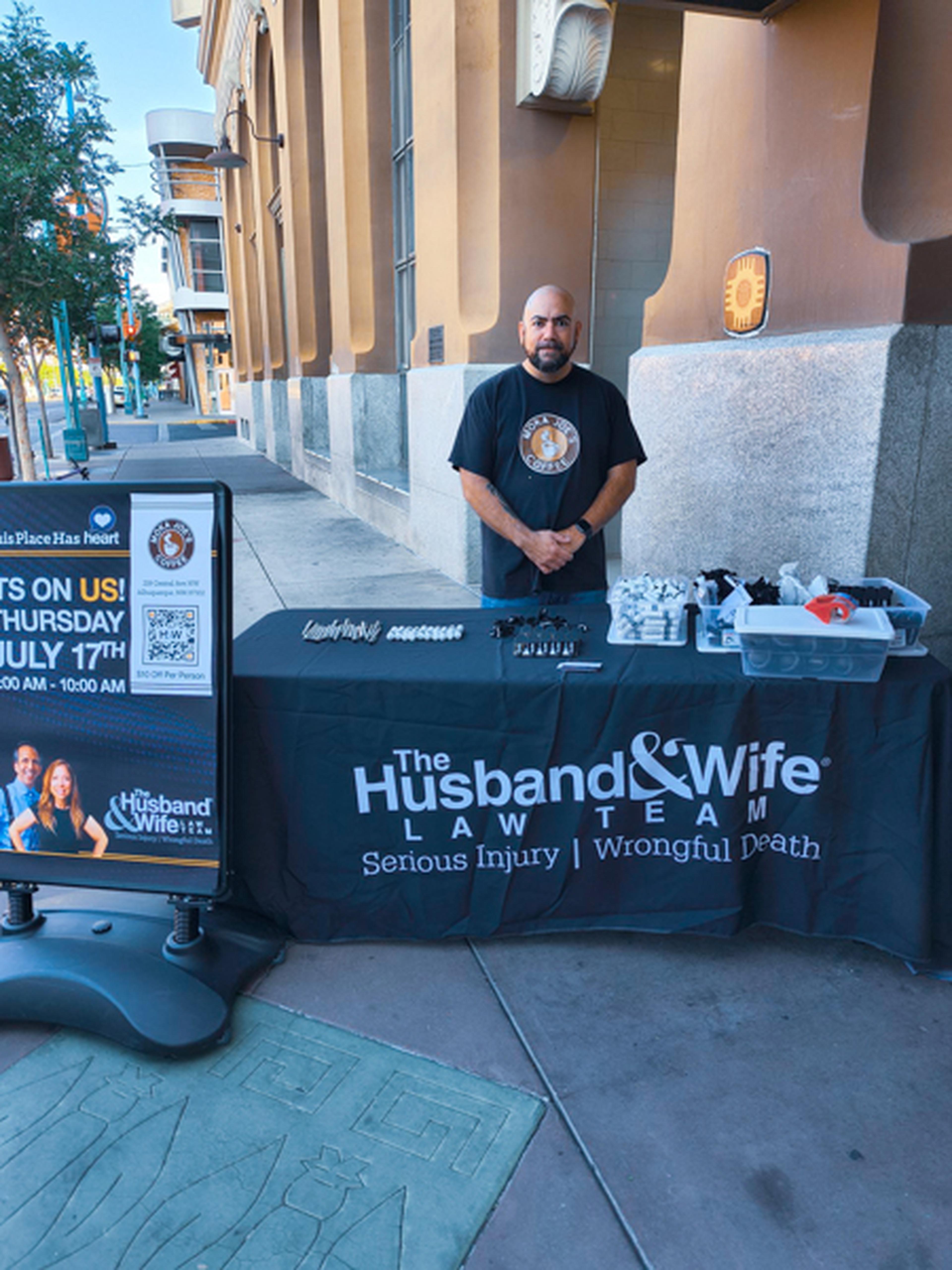 A man stands behind The Husband & Wife Law Team's branded table with giveaway items. The man is the owner of Moka Joe's