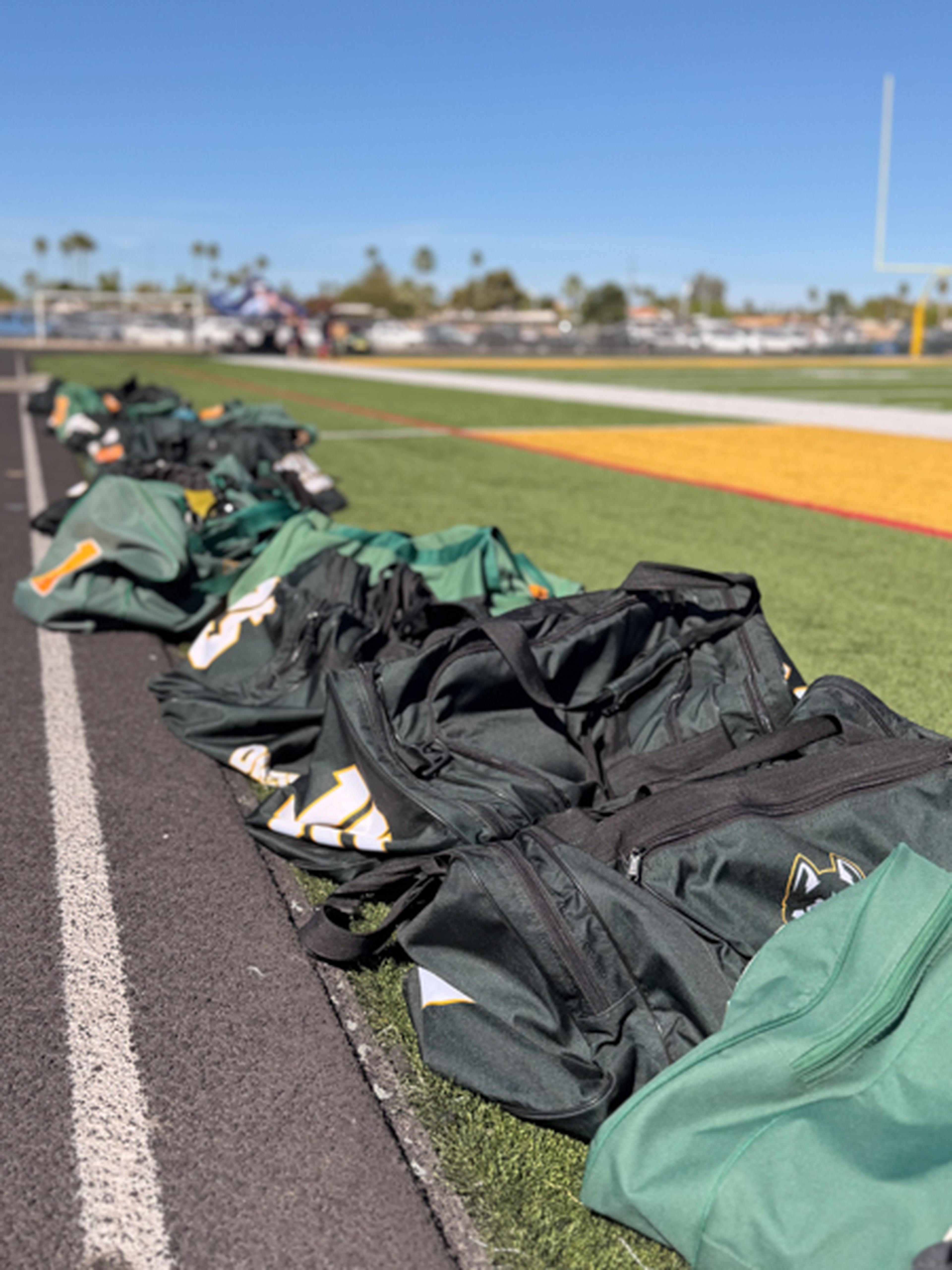 On a bright AZ field, green sports bags sit along the sideline beneath a clear sky and a visible goalpost.