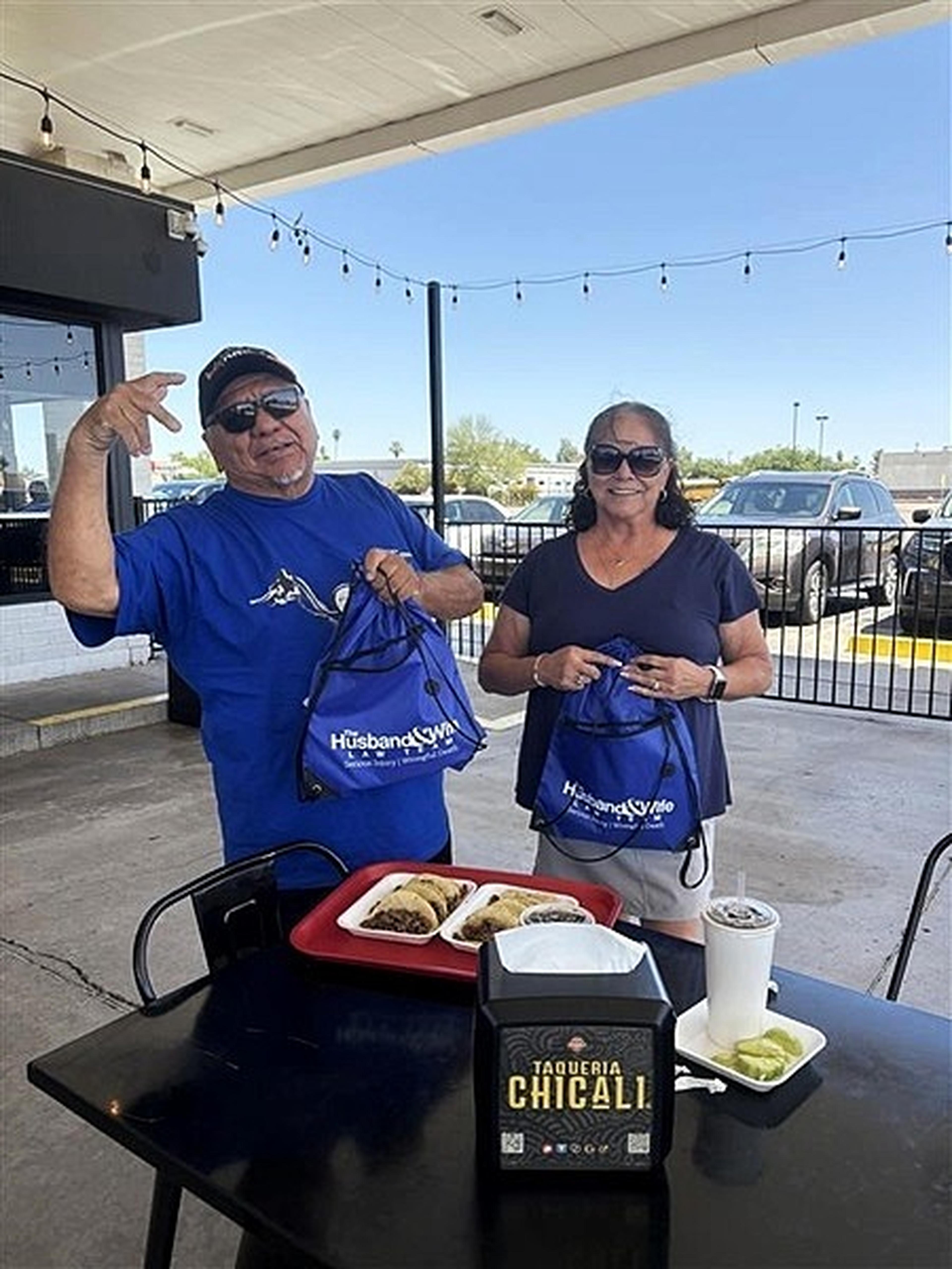 A smiling couple in matching shirts stands outside Taqueria Chicali holding bags, with a tray of tacos and a drink on the table.