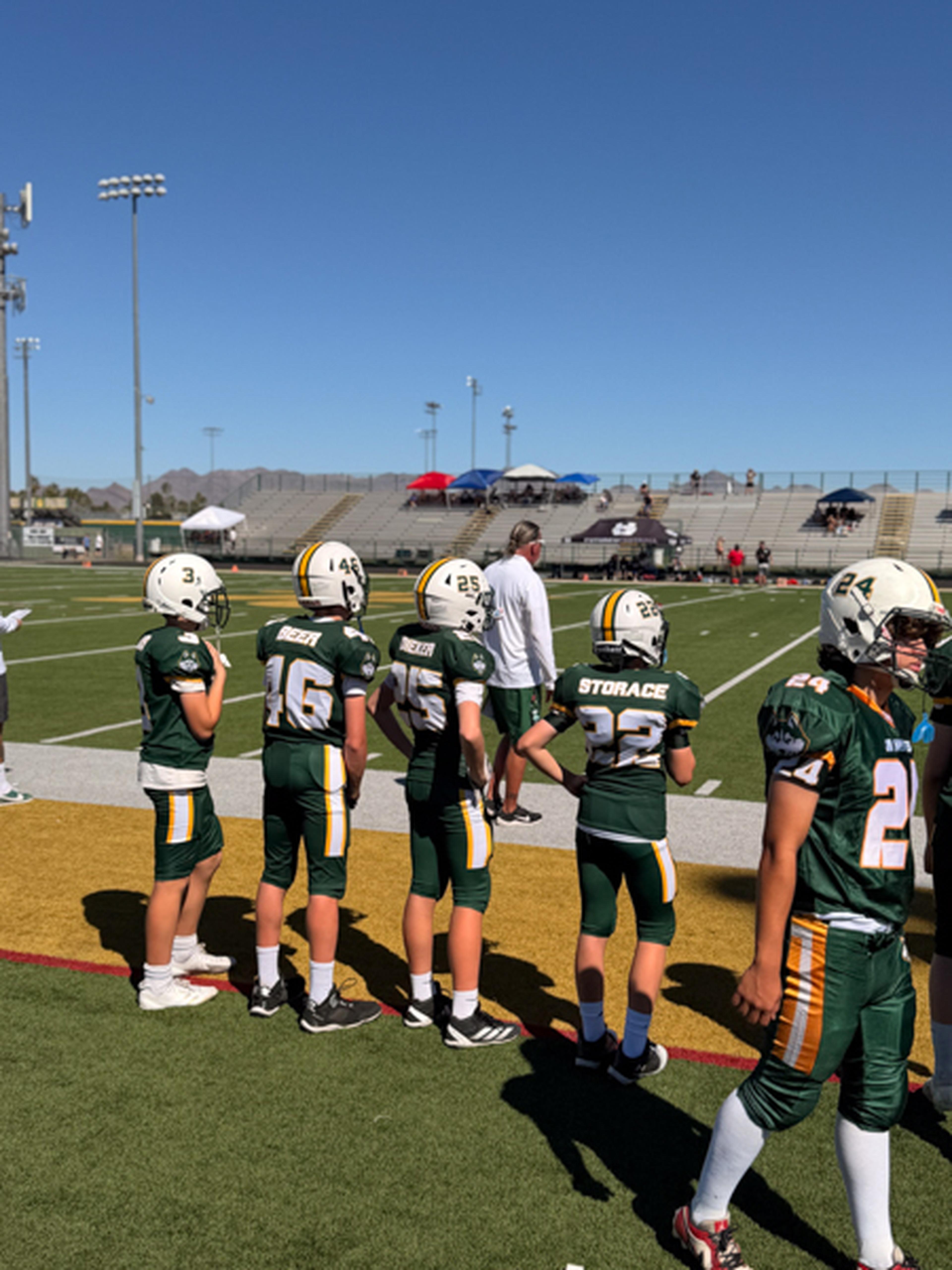 Youth football players in green uniforms stand on the sidelines with an empty stadium and the clear Arizona sky behind them.