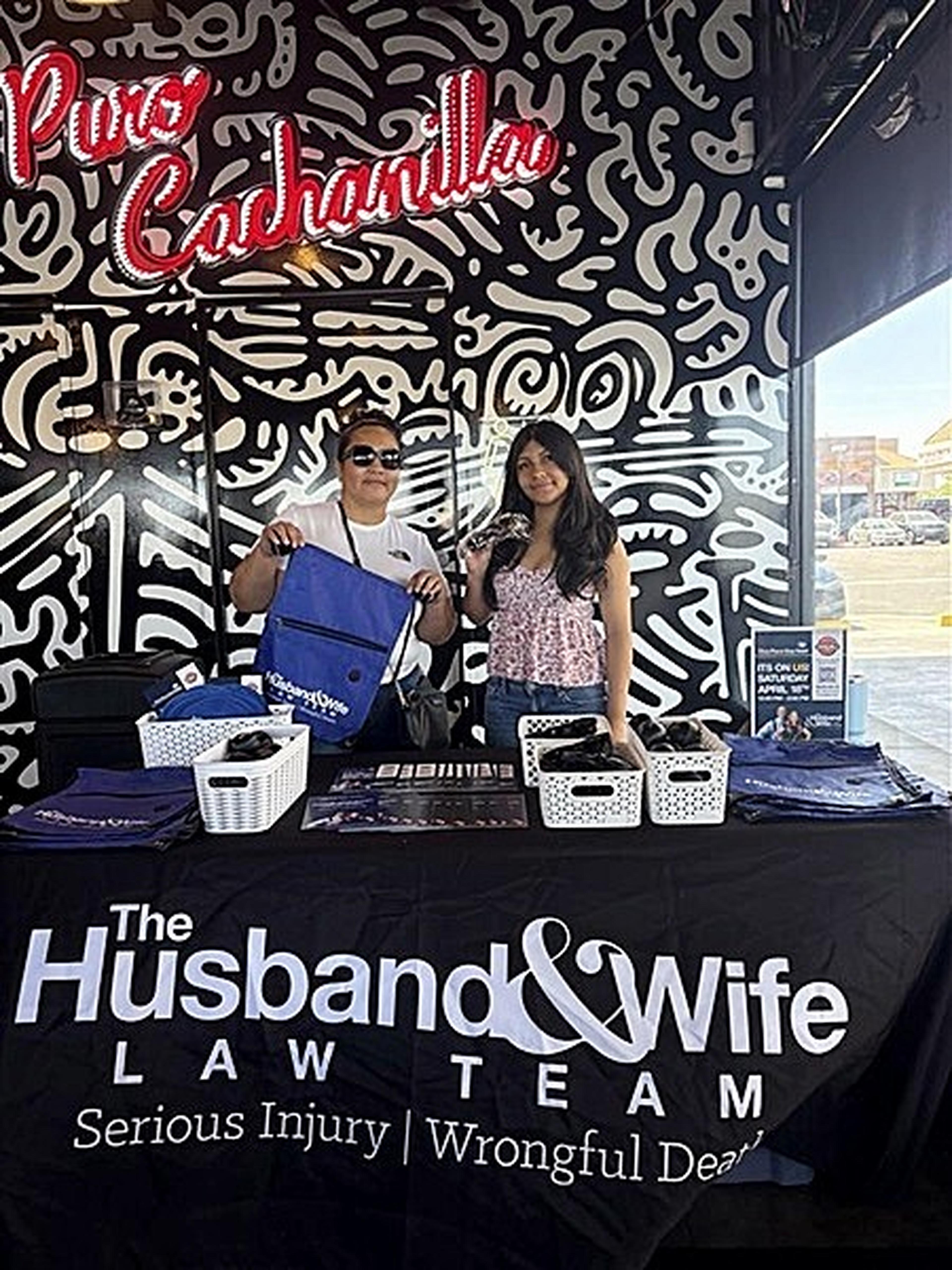 Two customers stand behind a Taqueria Chicali table branded with the Husband & Wife Law Team, holding promotional items.