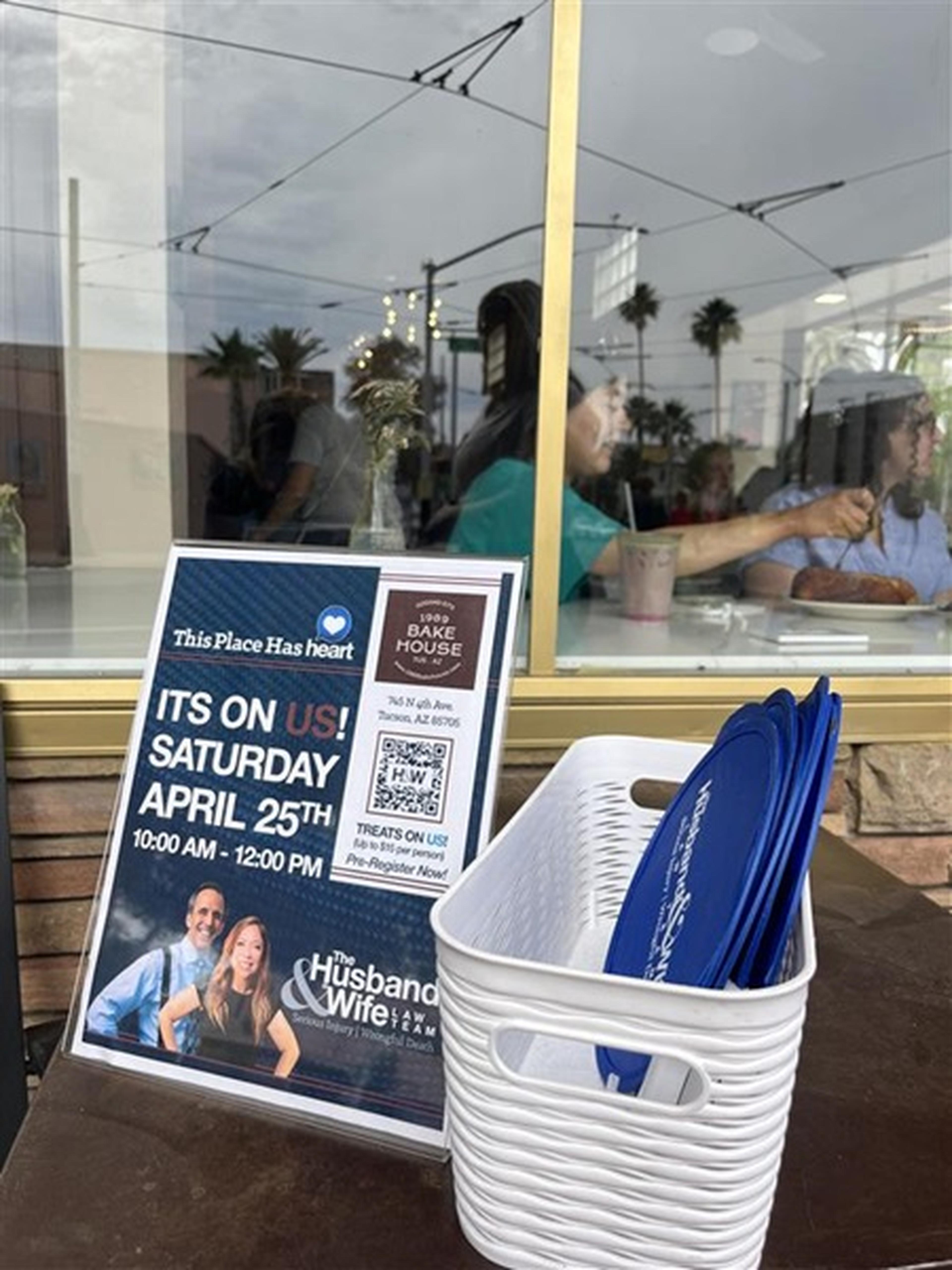 A flyer for the Husband & Wife Law Team event on April 25th sits beside a basket of blue fans, with people visible through the café window.
