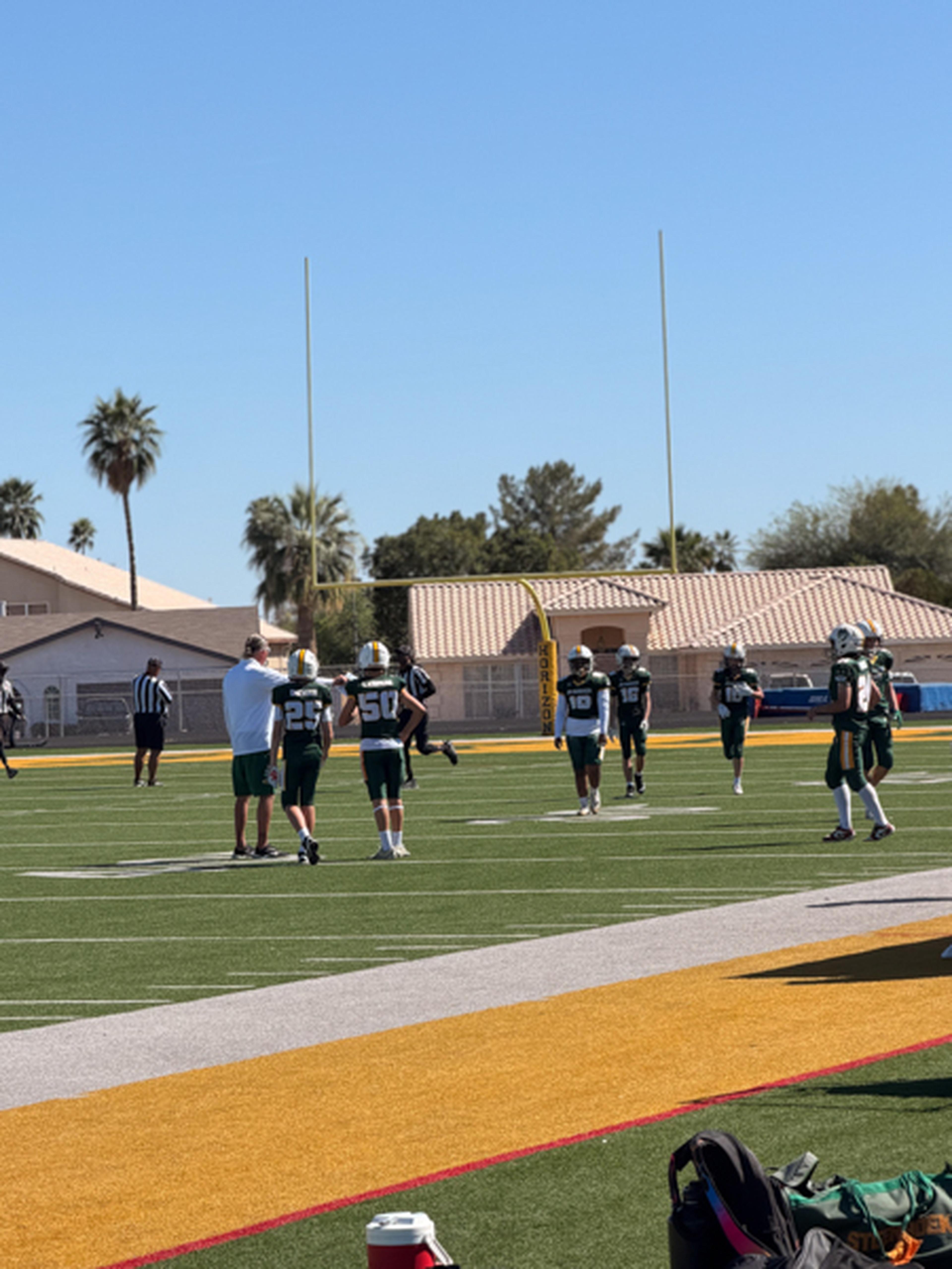 Coach Tomas and his players in green uniforms stand on a sunny field near the goalposts, getting ready for the game with palm trees and buildings in the background.