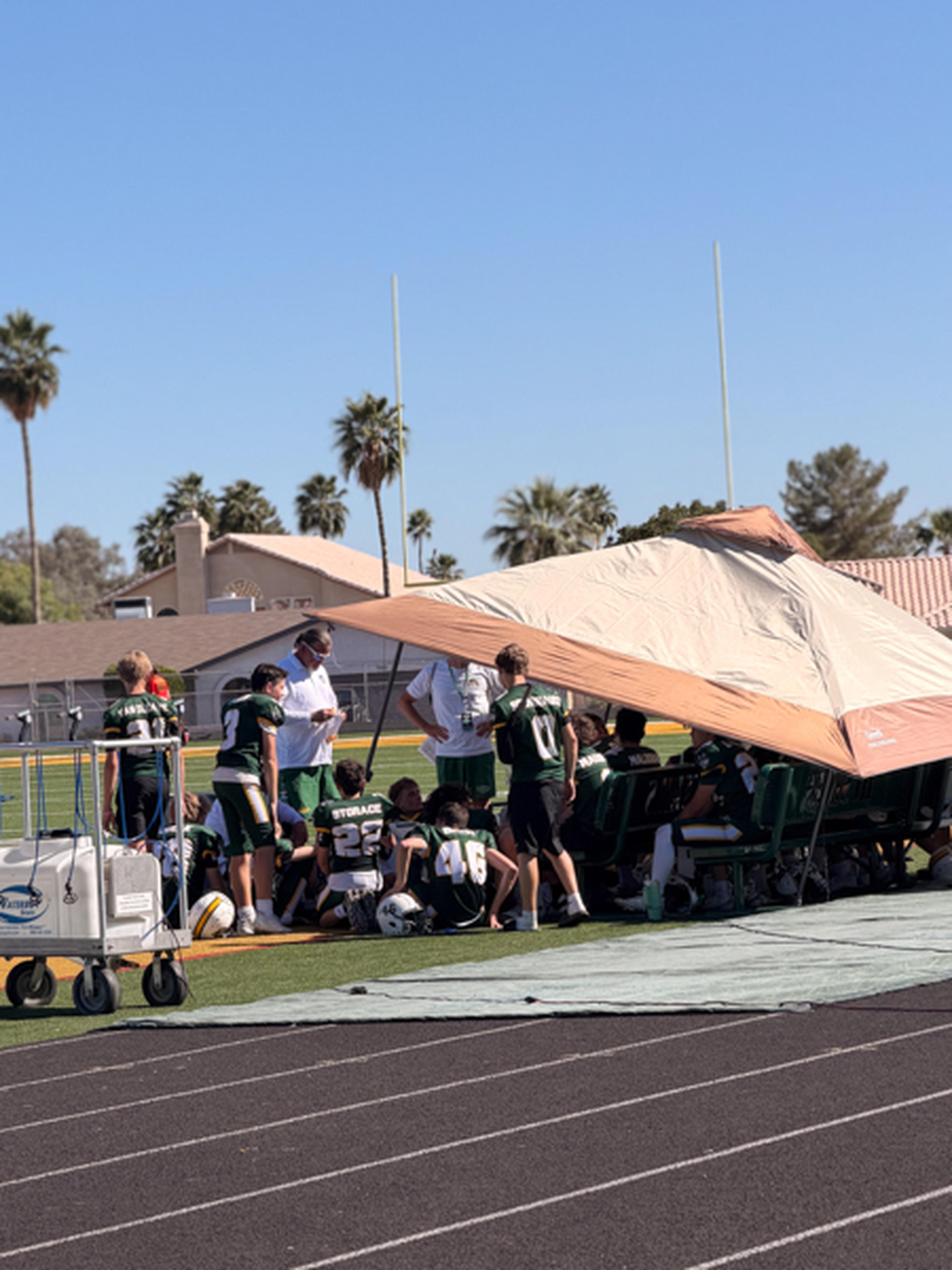 Arizona youth football team under a tent on the sidelines, with players resting and coaches standing, set against clear blue sky and palm trees.