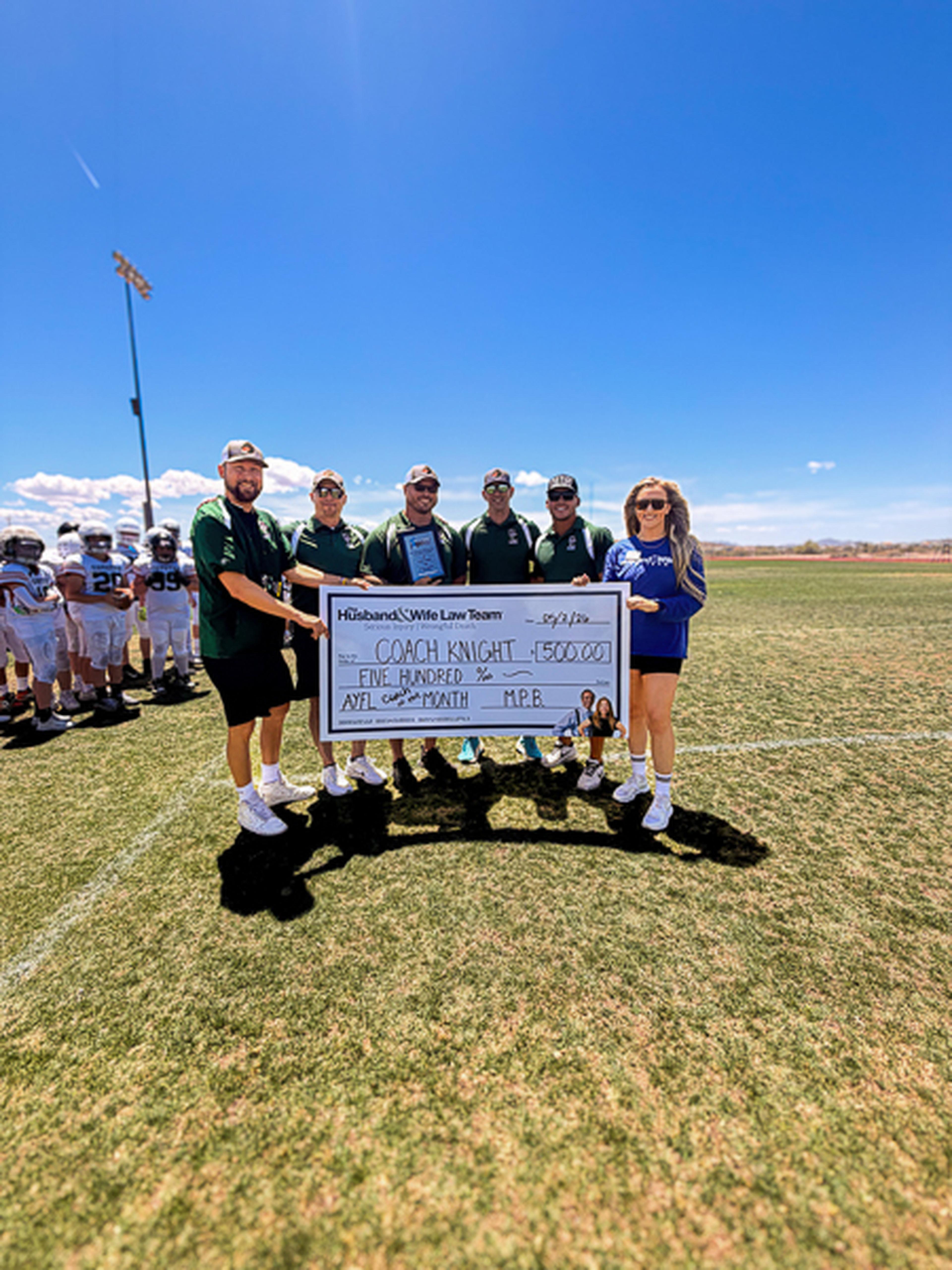 Football coaches gather on the field holding a $500 ceremonial check, with young players practicing behind them
