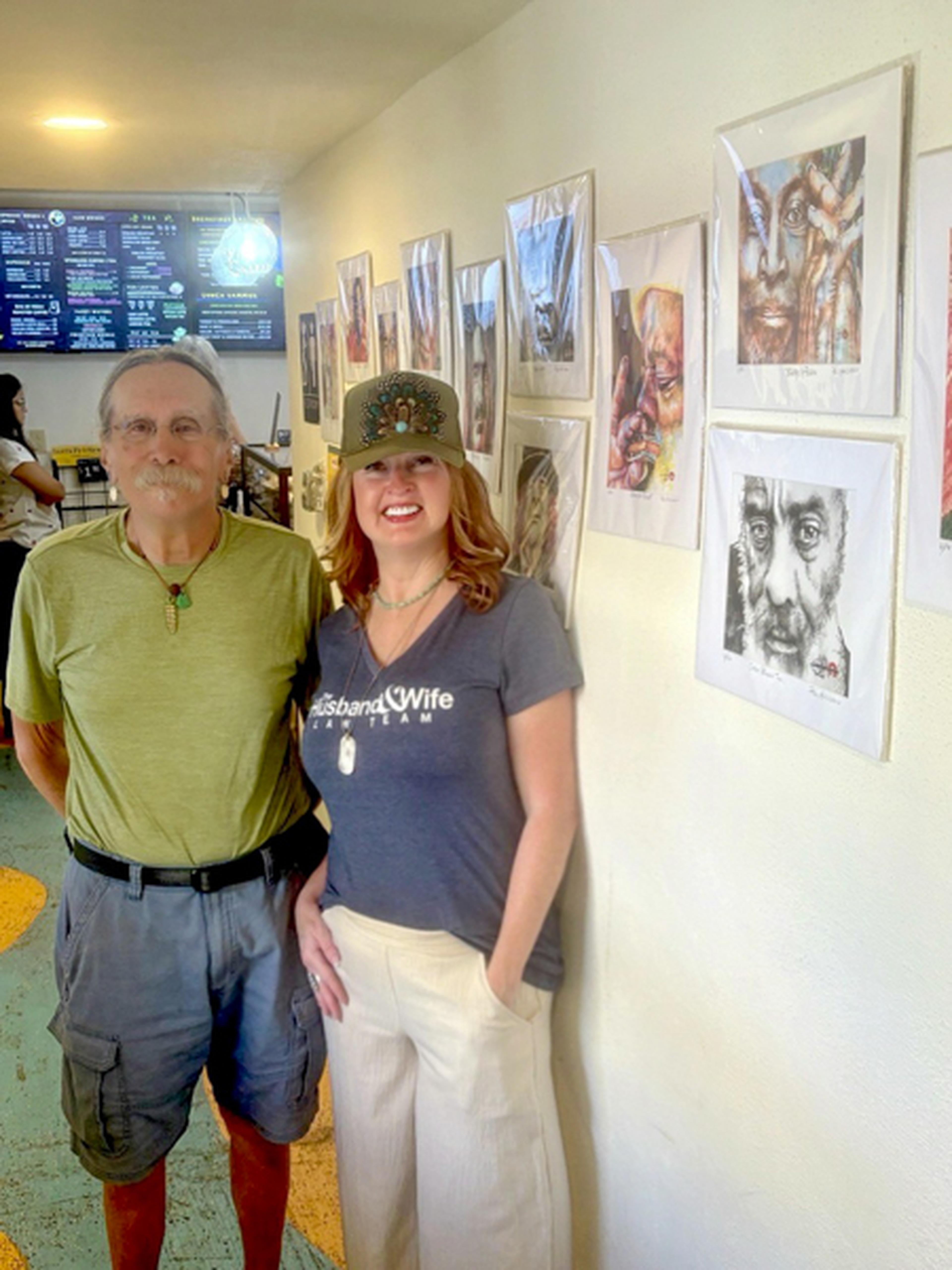 Two people stand smiling in an art gallery, with various portraits displayed on the wall behind them.
