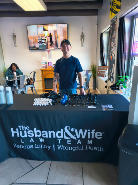 Shake Lab + The Husband & Wife Law Team are ready for the event! Man standing behind a table with promotional items for "The Husband & Wife Law Team" in a cafe setting.