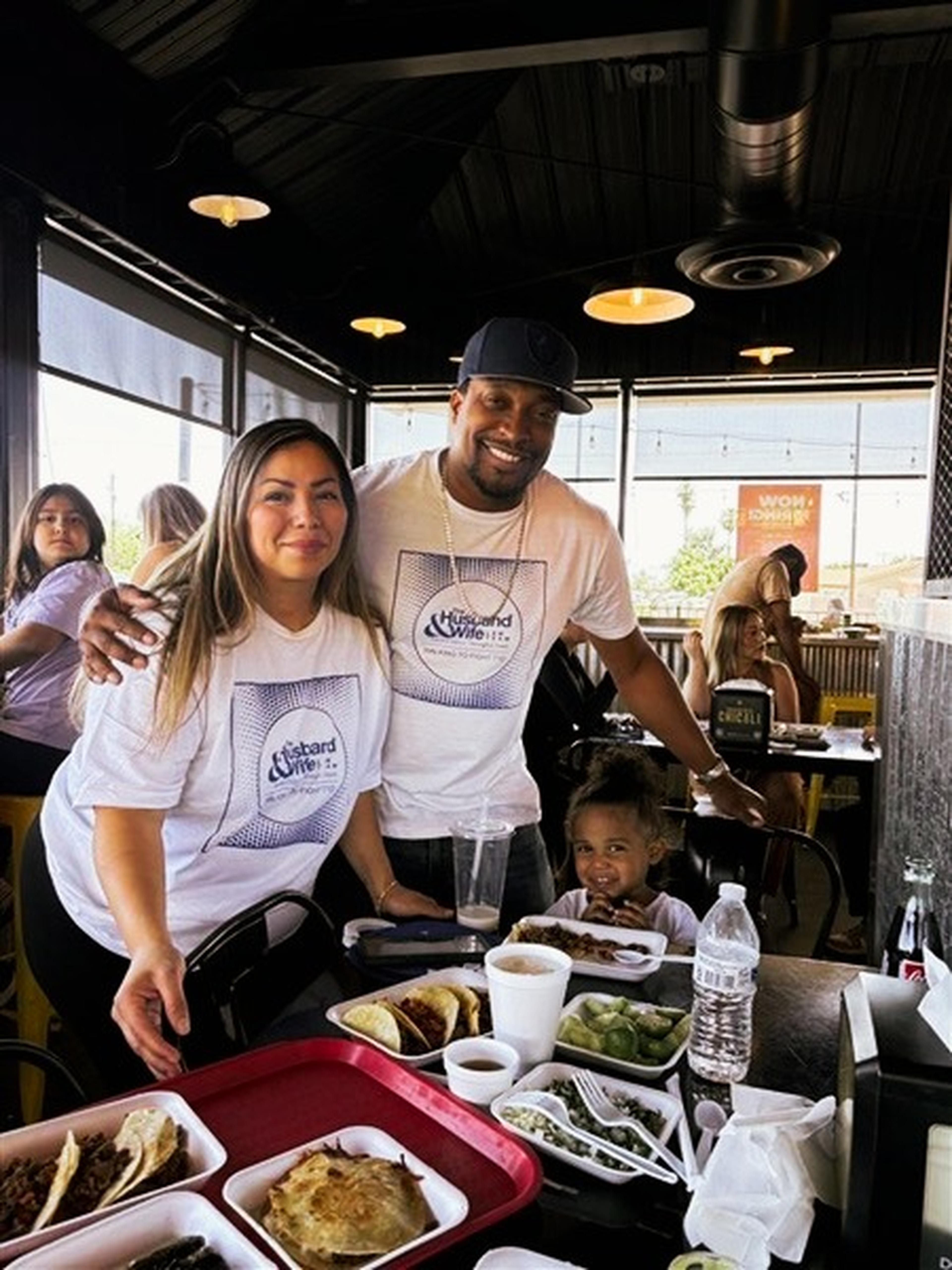 A smiling couple and their child sit at a table inside Taqueria Chicali with assorted dishes, all wearing matching event shirts.
