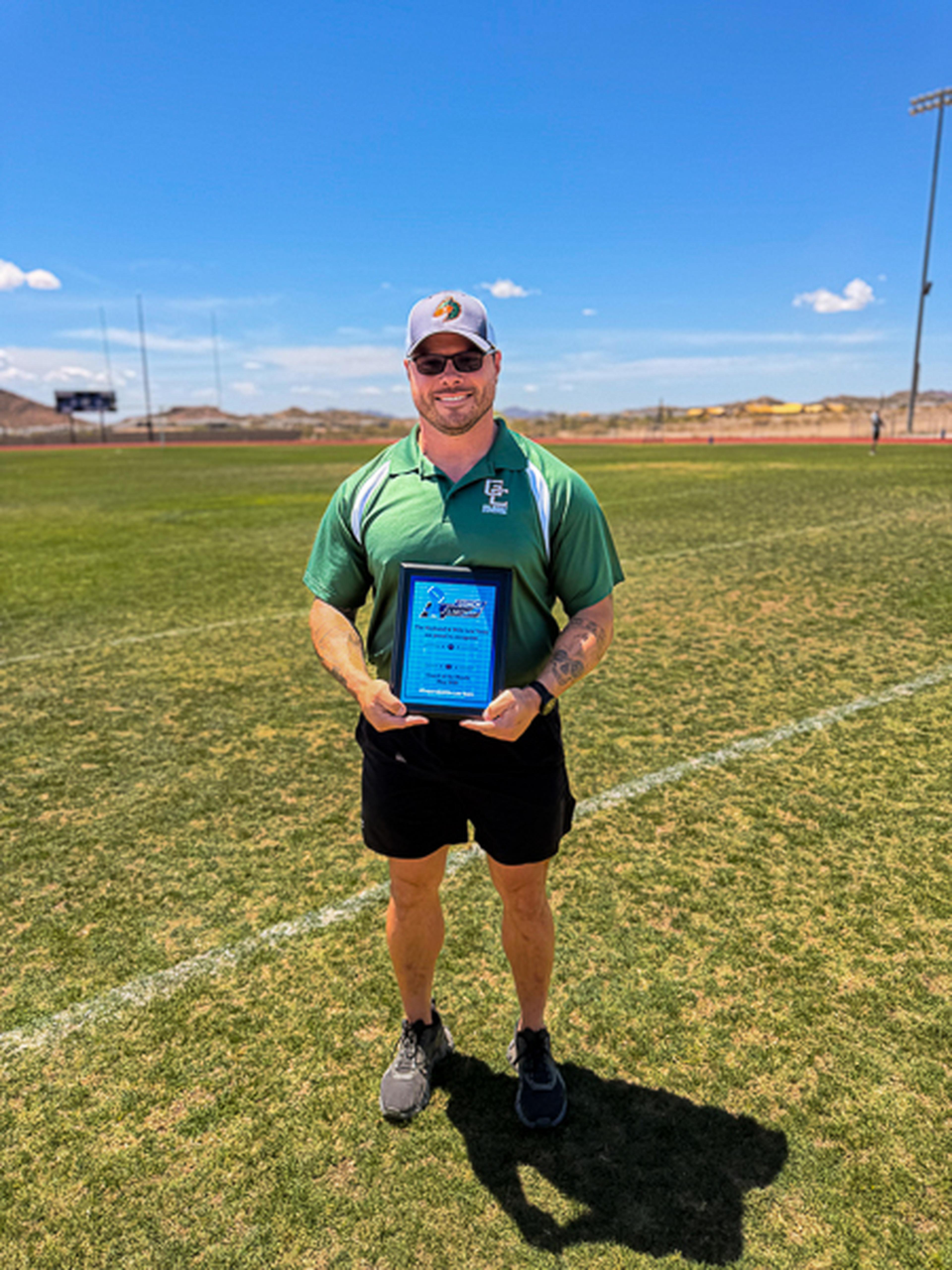 In a green shirt and cap, Coach Knight stands on the field proudly holding his award beneath a bright blue sky.