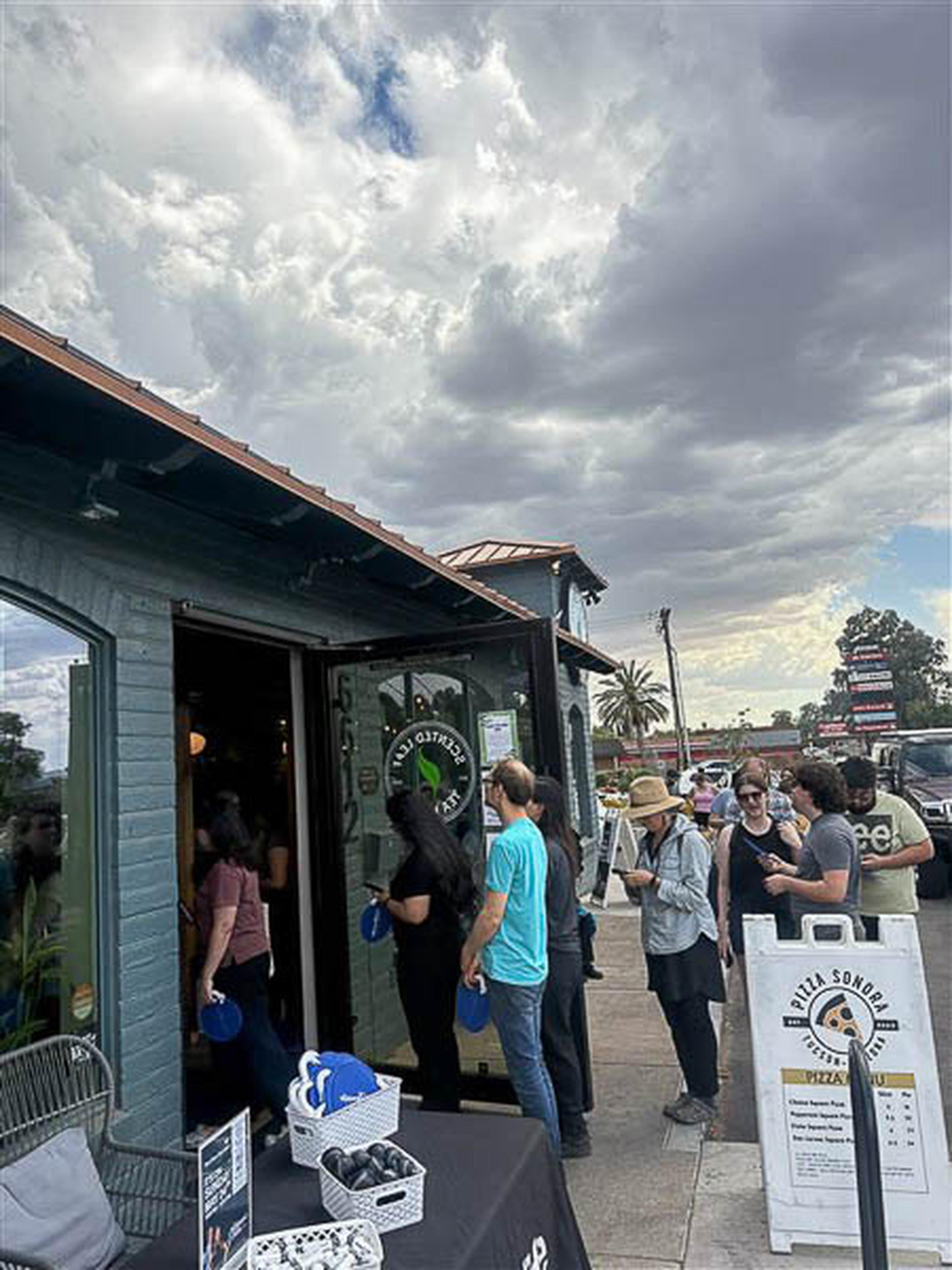 A line of customers waits outside Scented Leaf Tea House beneath a cloudy sky, with signs and outdoor tables in view.