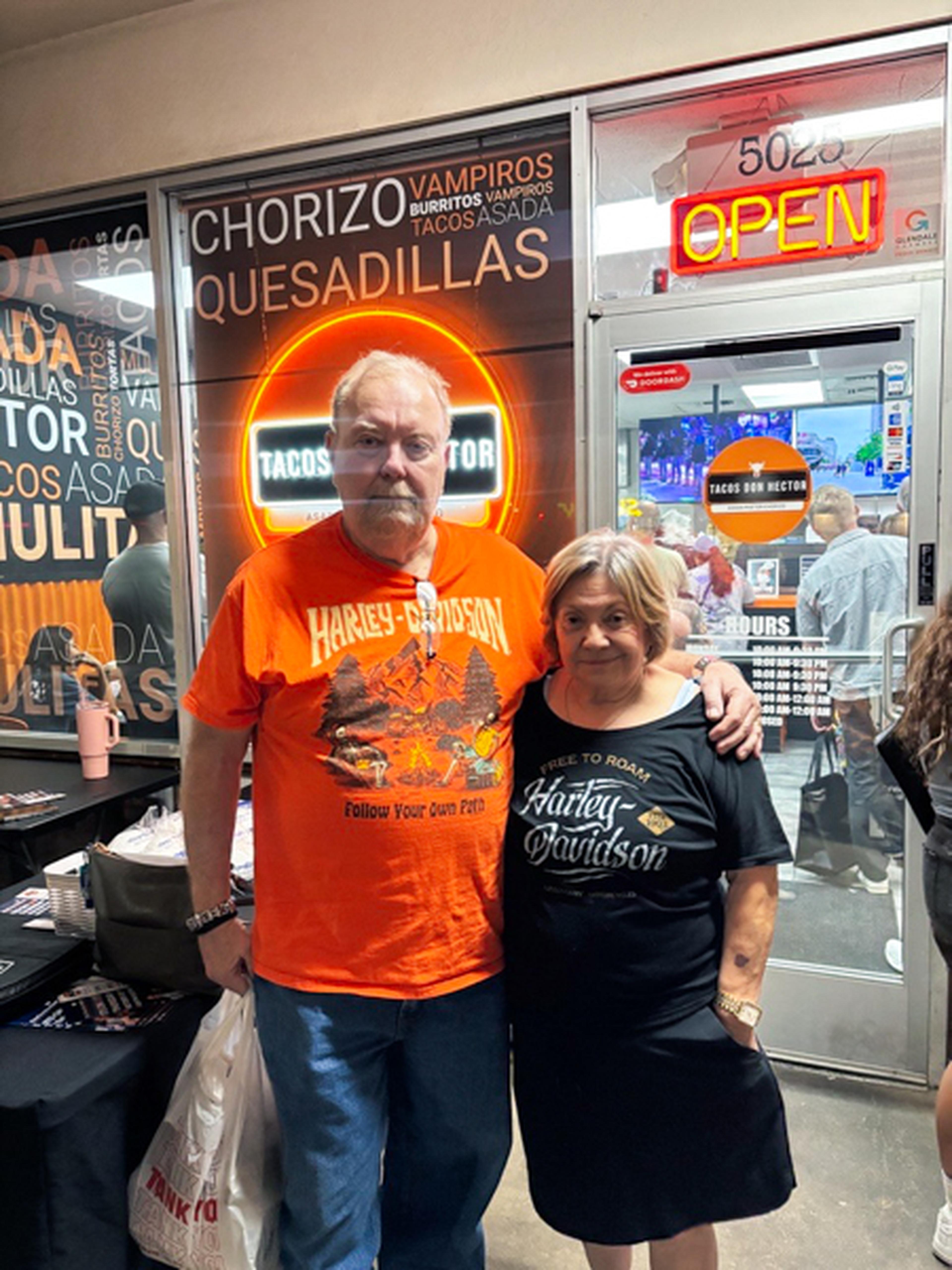 A man and woman stand together in front of a taco shop with neon signs and an open door.